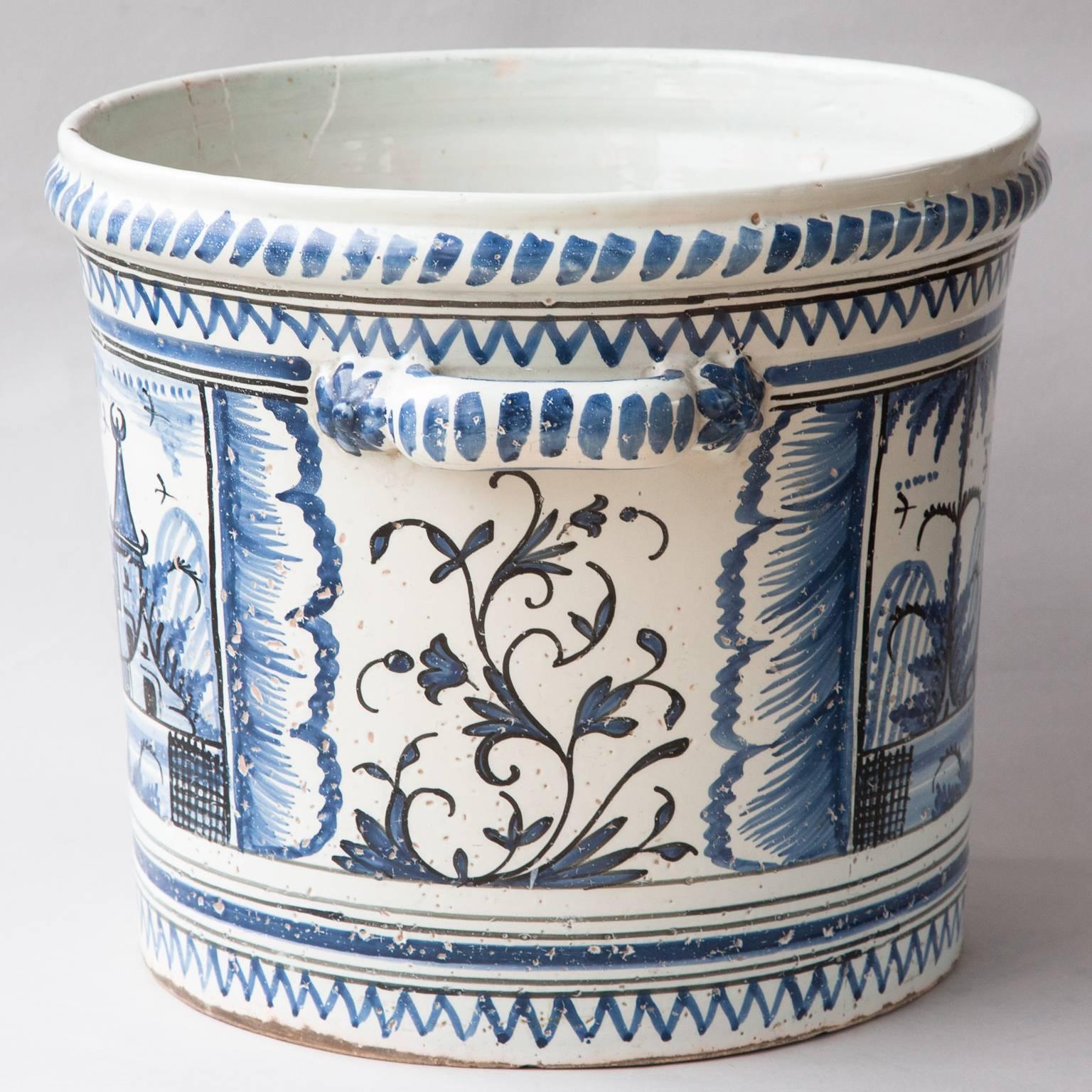 Decorated in cobalt blue on a milky white ground with scenes of buildings in landscapes in panels, with zig zag decoration circling the pot at the bottom and under the upper rim. The two handles with blue flowers on either side. 
France, circa