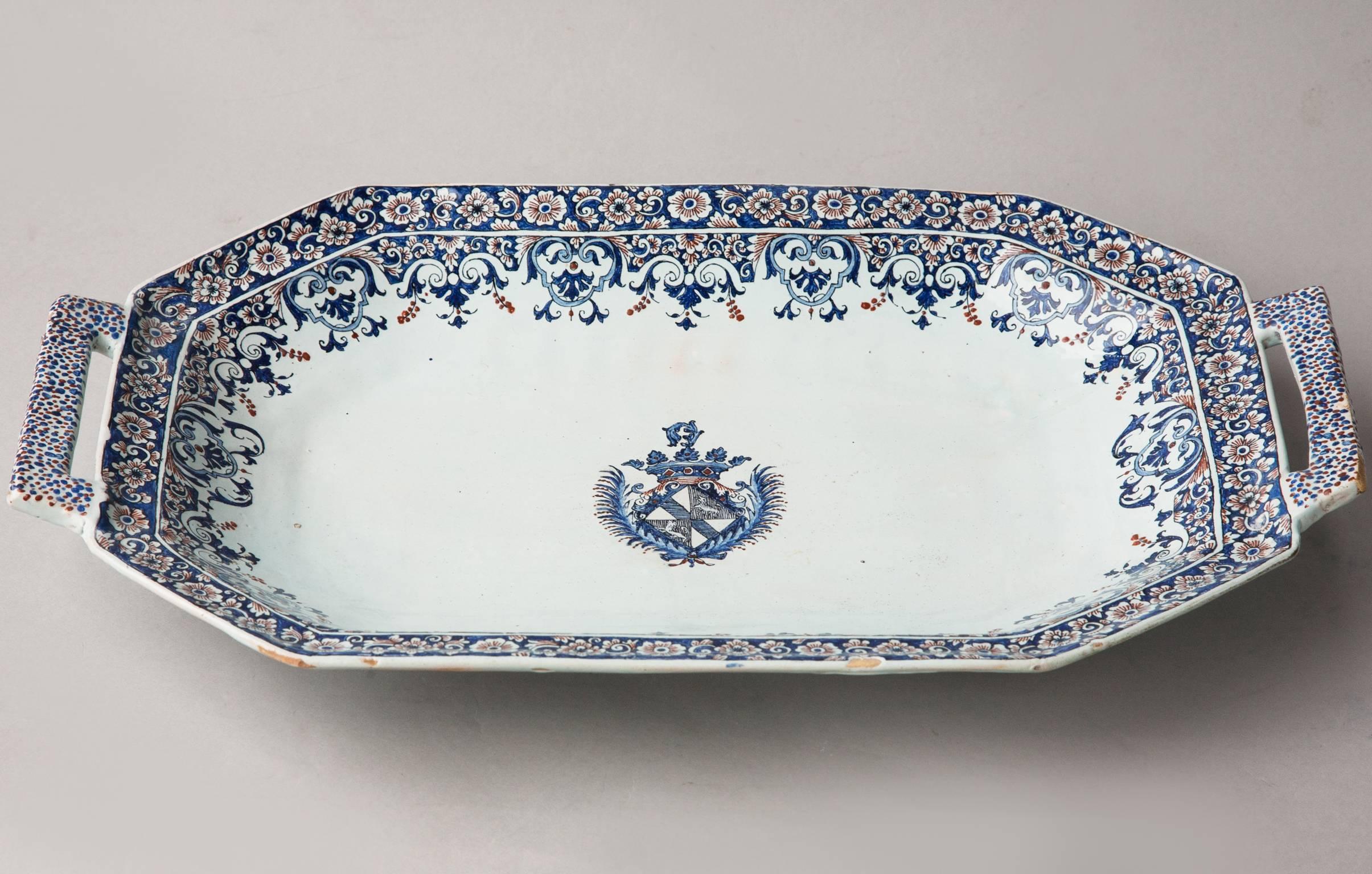 Decorated with a geometric cobalt blue and red floral border with a coat of arms at the centre. With two handles, one at each end. 
Rouen, France, circa 1710.
A small amount of restoration to the underside of the handles. 
There is a dish with