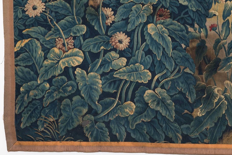 Verdure tapestry with a large house in the distance and two stags being chased by two hounds. . 
The foreground has a rich floral and foliate decoration with poppies, flowers and fruit trees. Surrounded by a plain rust coloured border.
Finely