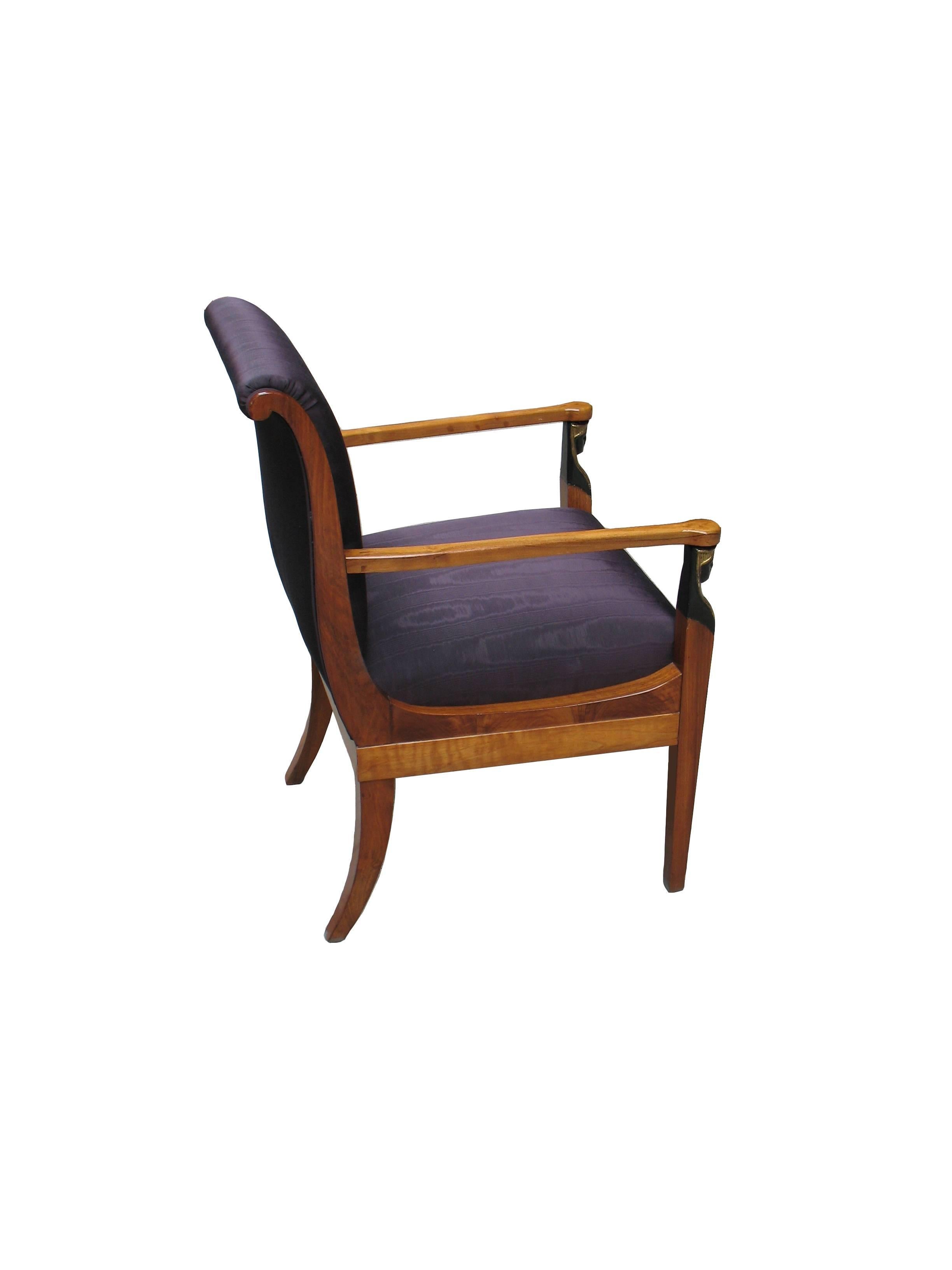 This South German Biedermeier armchair, exemplary designed with straight tail, arms, frontlegs, outbent padded seatback and saber legs, features ebonized and parcel - gilt Egyptian uprights and verifies the Classical Antiquity inspirations into the