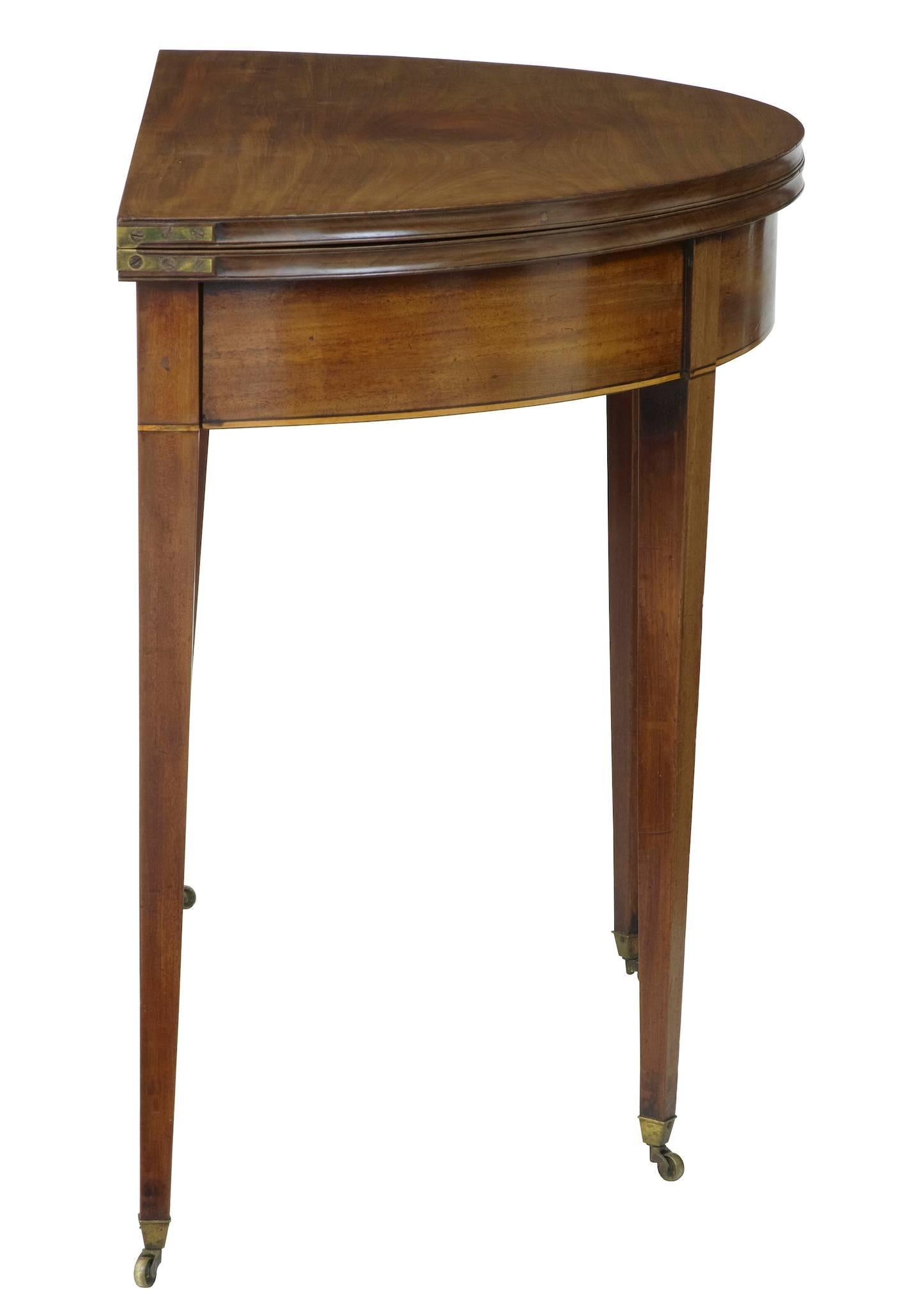 Mahogany card table, circa 1830.
demilune shaped with satinwood and ebony stringing to front.
Original color on the interior.
Standing on four tapering legs with brass castors.

Height: 28 3/4