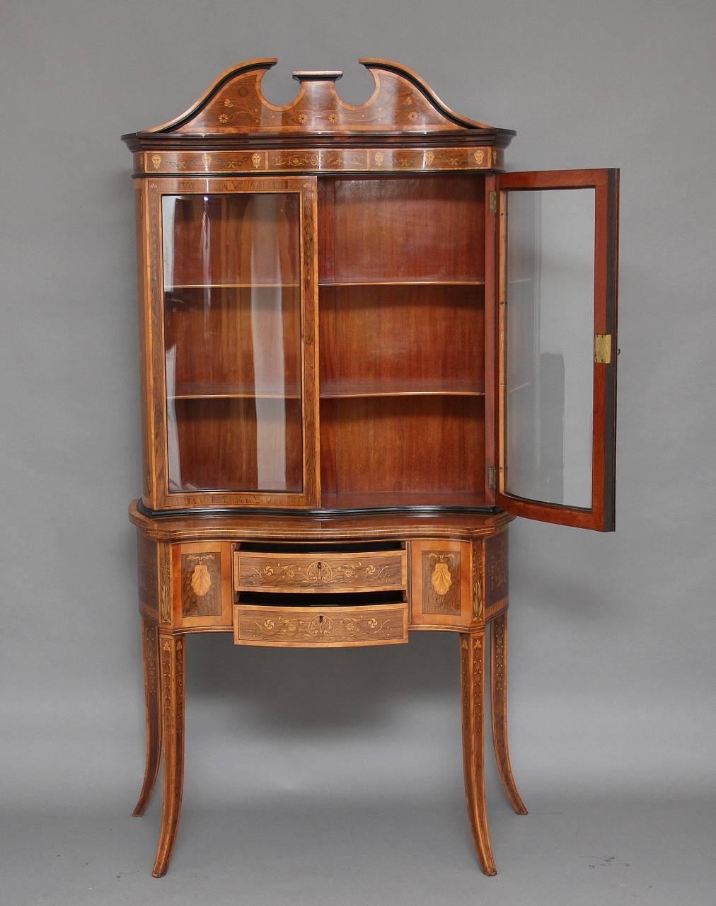 19th century Sheraton Revival rosewood display cabinet profusely inlaid all-over and crossbanded in satinwood, the top having a broken arch pediment and below the frieze decorated with inlaid male masks, with serpentine shaped doors and glass