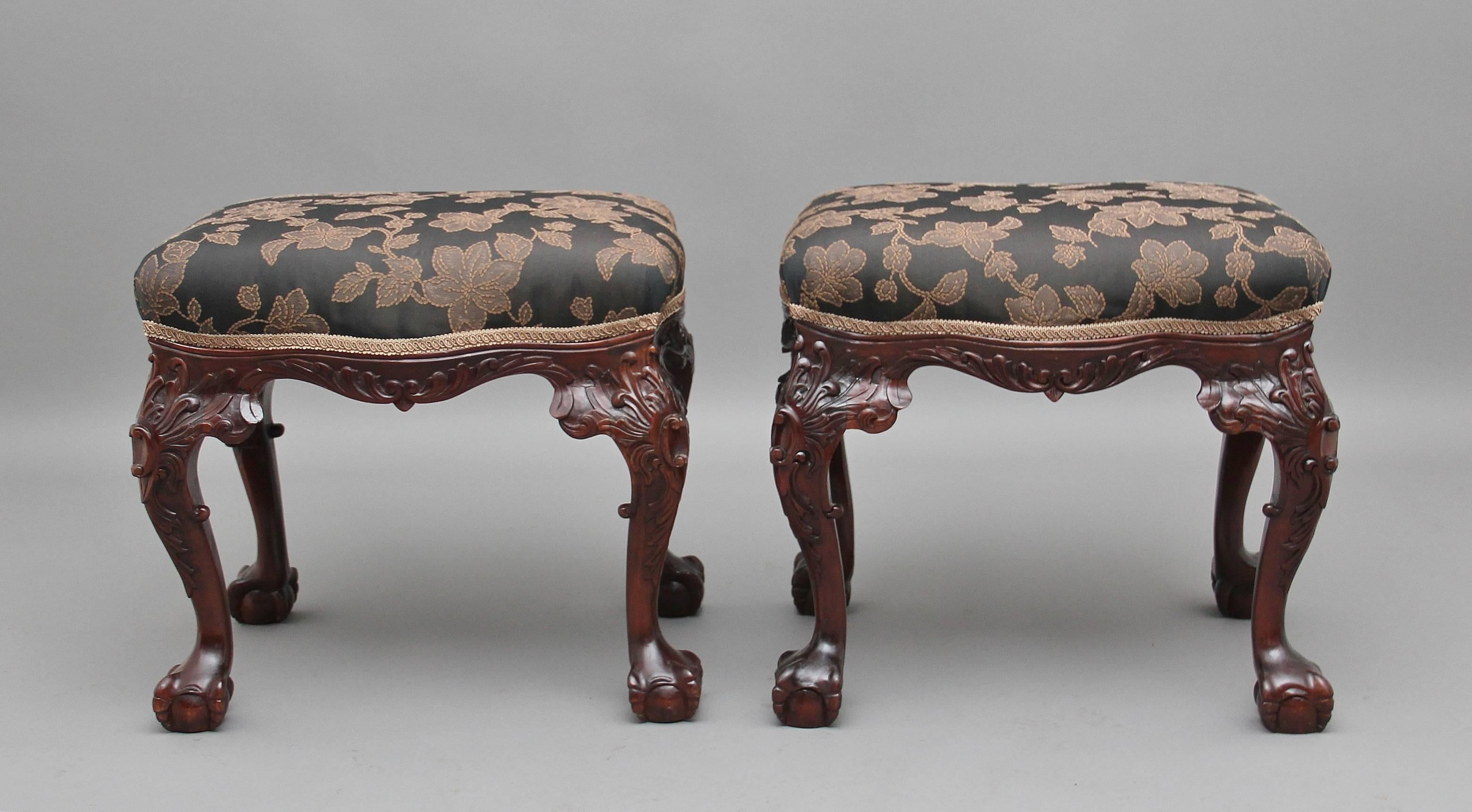 A decorative pair of mid-20th century mahogany stools, each of serpentine outline, upholstered in a dark colored fabric with a floral pattern, supported on foliate carved cabriole legs terminating in claw and ball feet, circa 1950. Measures: Height