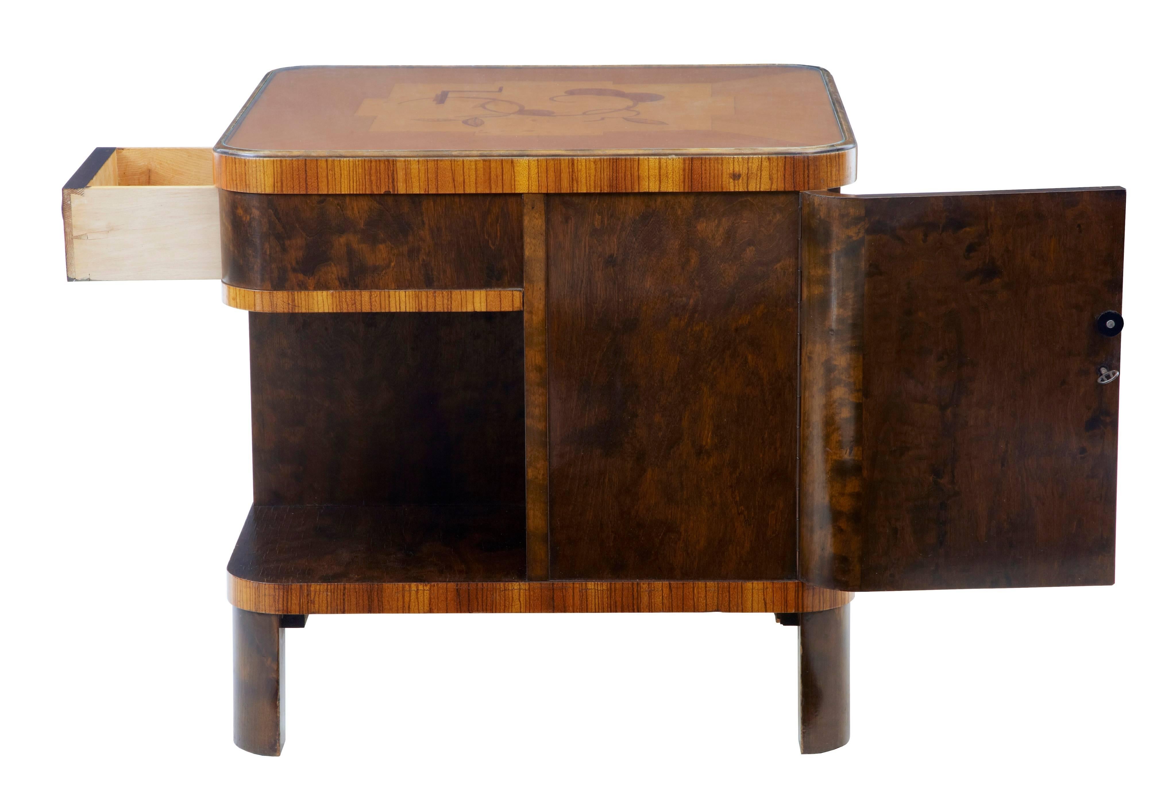 Good quality two-tone birch occasional table, circa 1930.
Rounded square in shape with a drawer, single door cupboard and open storage spaces.
Inlaid top with removable glass protection.

Measures: Height: 23