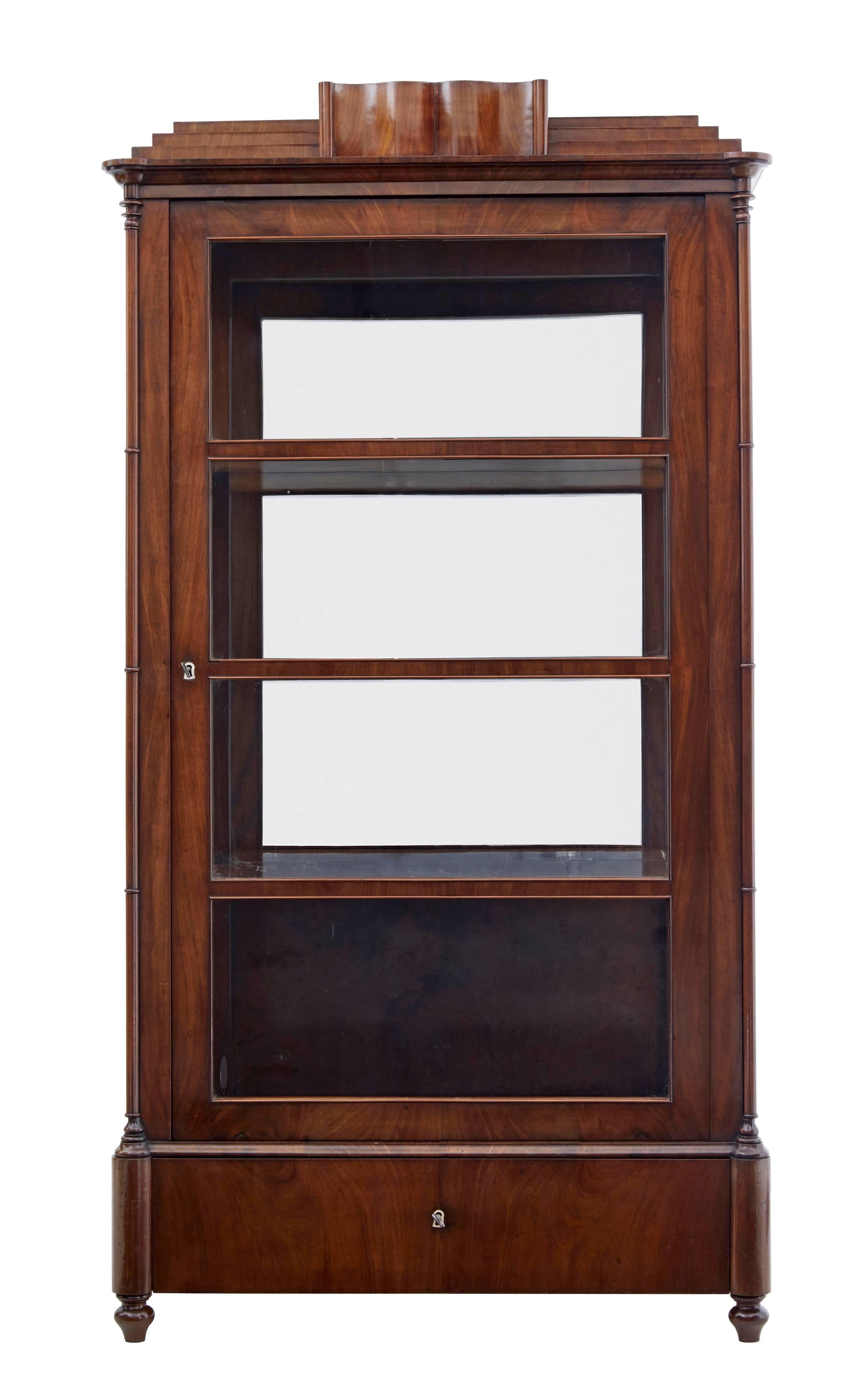 Rare Danish flame mahogany display cabinet, circa 1840.
Single door cabinet containing three mahogany shelves with original mirrored back panels.
Original glazing to the front and sides.
Concealed drawer in the pediment with a further deep drawer