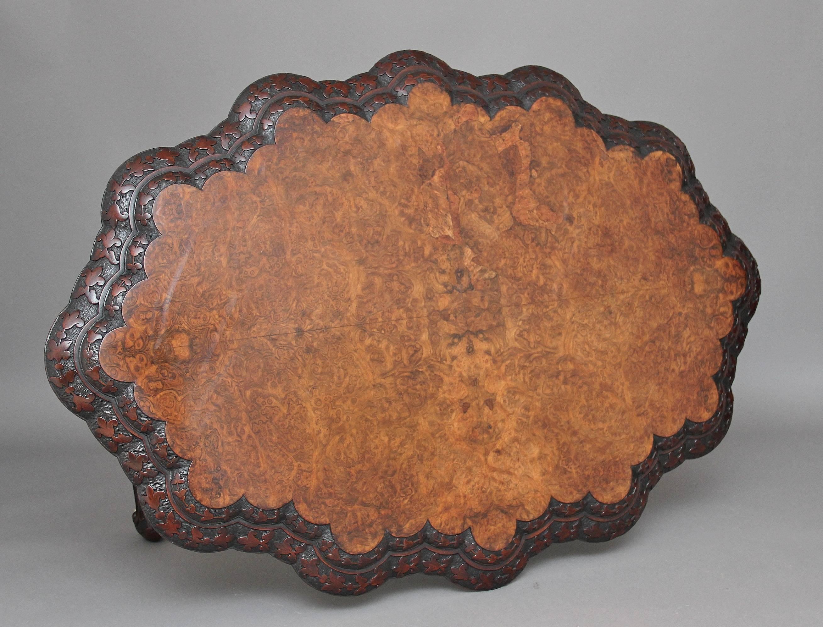 19th century Irish burr walnut stretcher or centre table, the scalloped shaped quarter veneered top having a double moulded and trailing leaf and flower carved edge, supported on baluster turned end supports with outswept legs terminating on ceramic