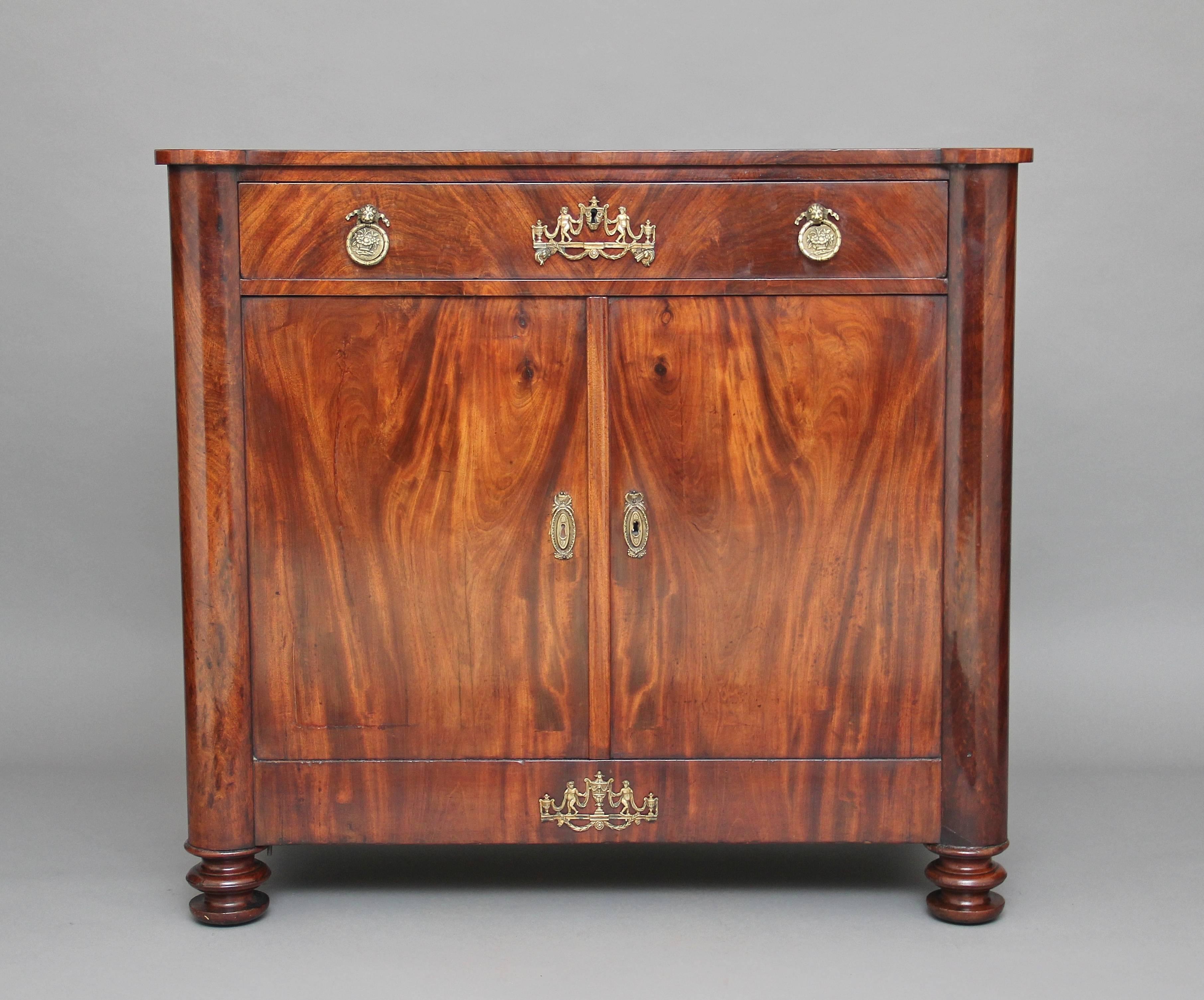 19th century French mahogany cabinet, having a single drawer above a two door cupboard with a single shelf inside, the drawer and doors having brass mask handles, oval escutcheons and applied decoration, flanked either side by half columns,