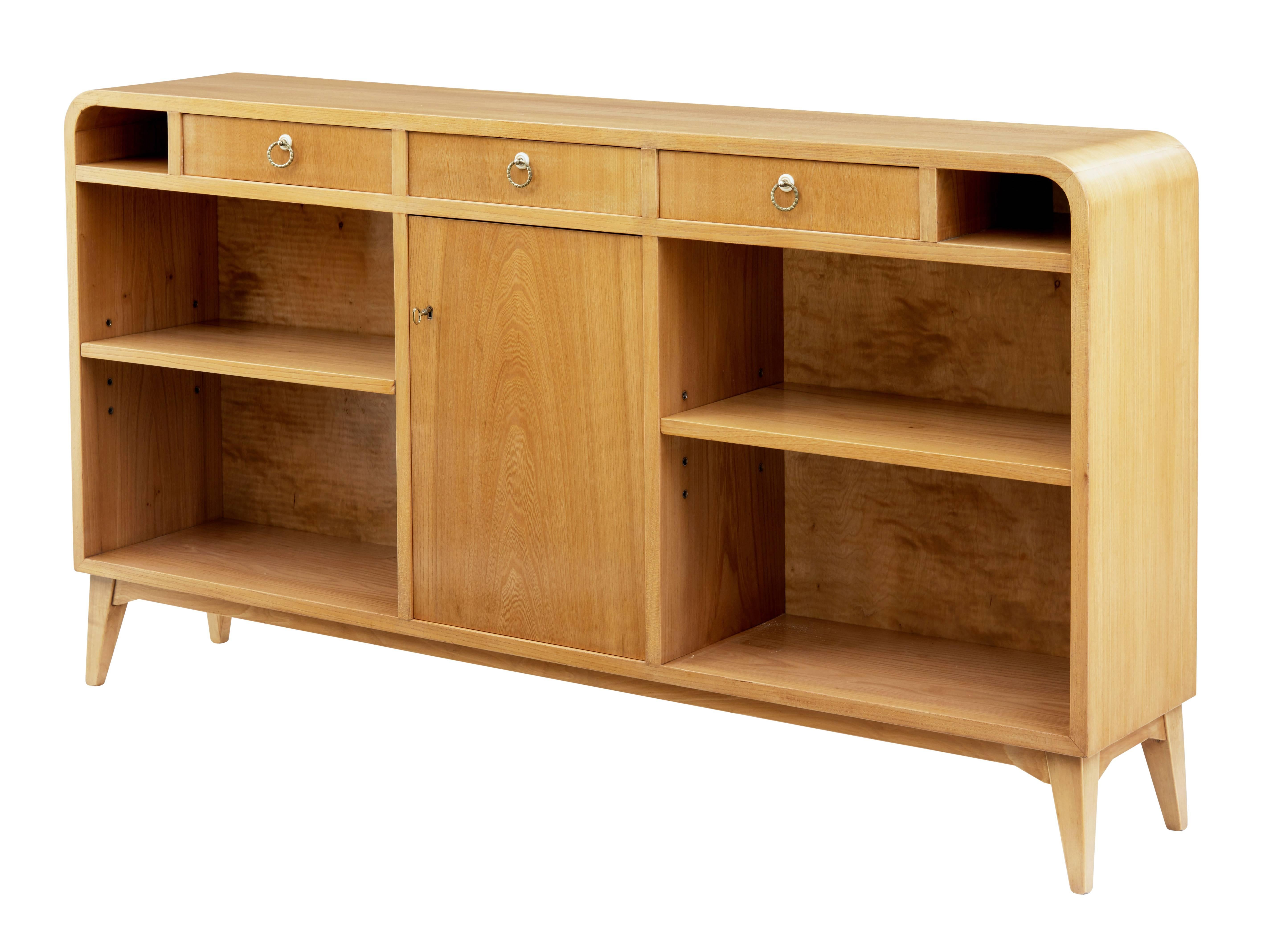 Excellent quality piece of Scandinavian 1950s design.

Three central drawers with an open storage aperture which follows the curved ends either end.

Central lockable single door with interior shelf, flanked either side by single shelf open