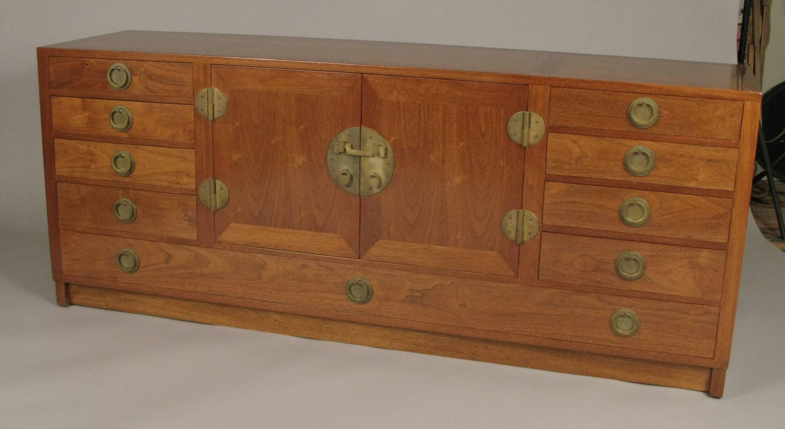 A beautiful and Classic 1950s walnut and mahogany sideboard designed by Edward Wormley for Dunbar, with solid brass hardware in an Asian Modern style. The centre section has a pair of doors concealing a glass shelf, and a removable glass serving
