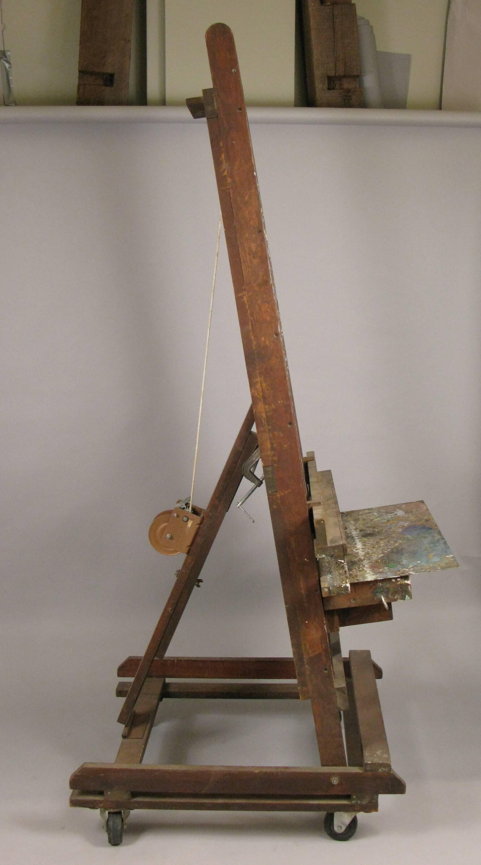 A very large-scale vintage 1950s adjustable artists easel, on wheels with a pulley and winch to raise or lower the large art shelf. In original condition with scattered paint.