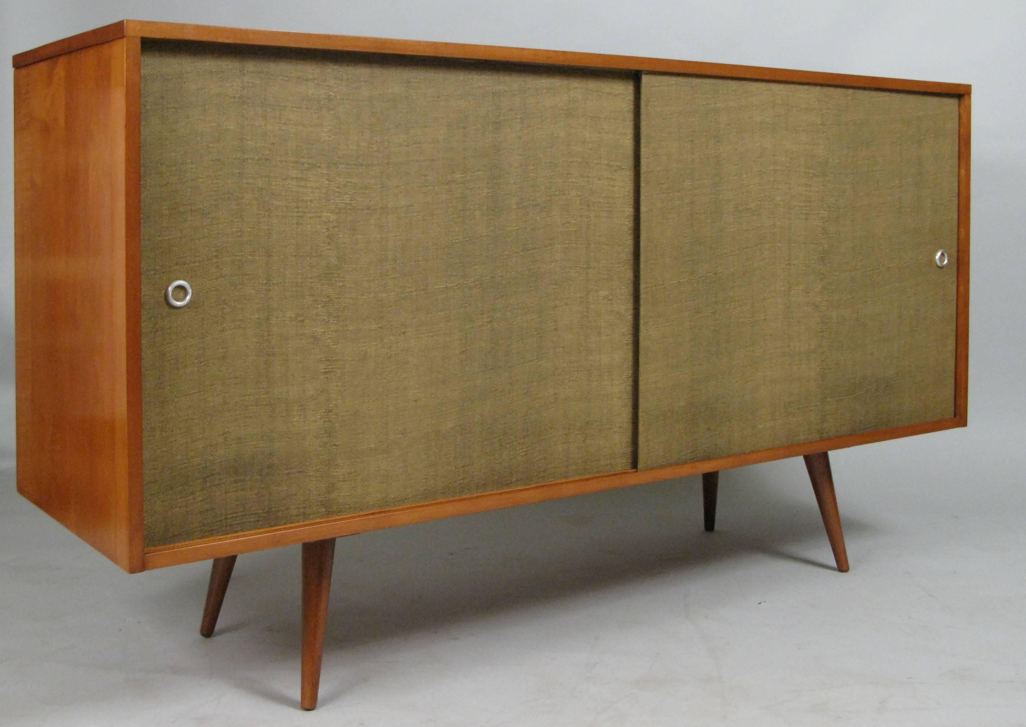 A classic mid-20th century birch credenza by Paul McCobb. Gracious large cabinet with sliding doors covered in McCobb's signature grasscloth with zinc hardware. Divided interior is fitted with four drawers and an adjustable shelf. Expertly restored