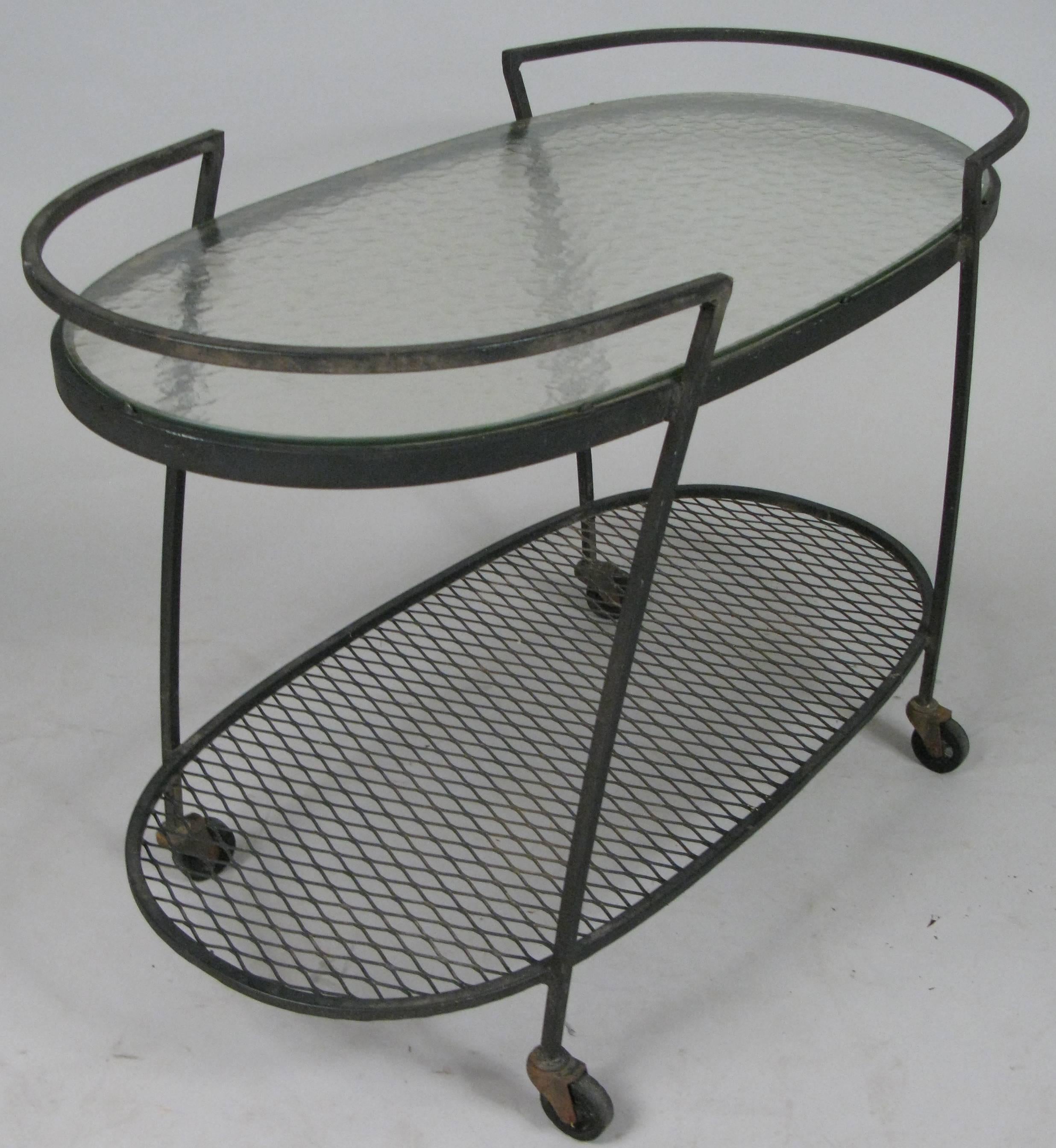 One of the best of Woodard's series of wrought iron rolling bar carts from the 1950s, this oval design has a low steel mesh shelf, and a large glass oval top. The handles curve around the ends and provide a rim to keep bottles and glass on the cart.