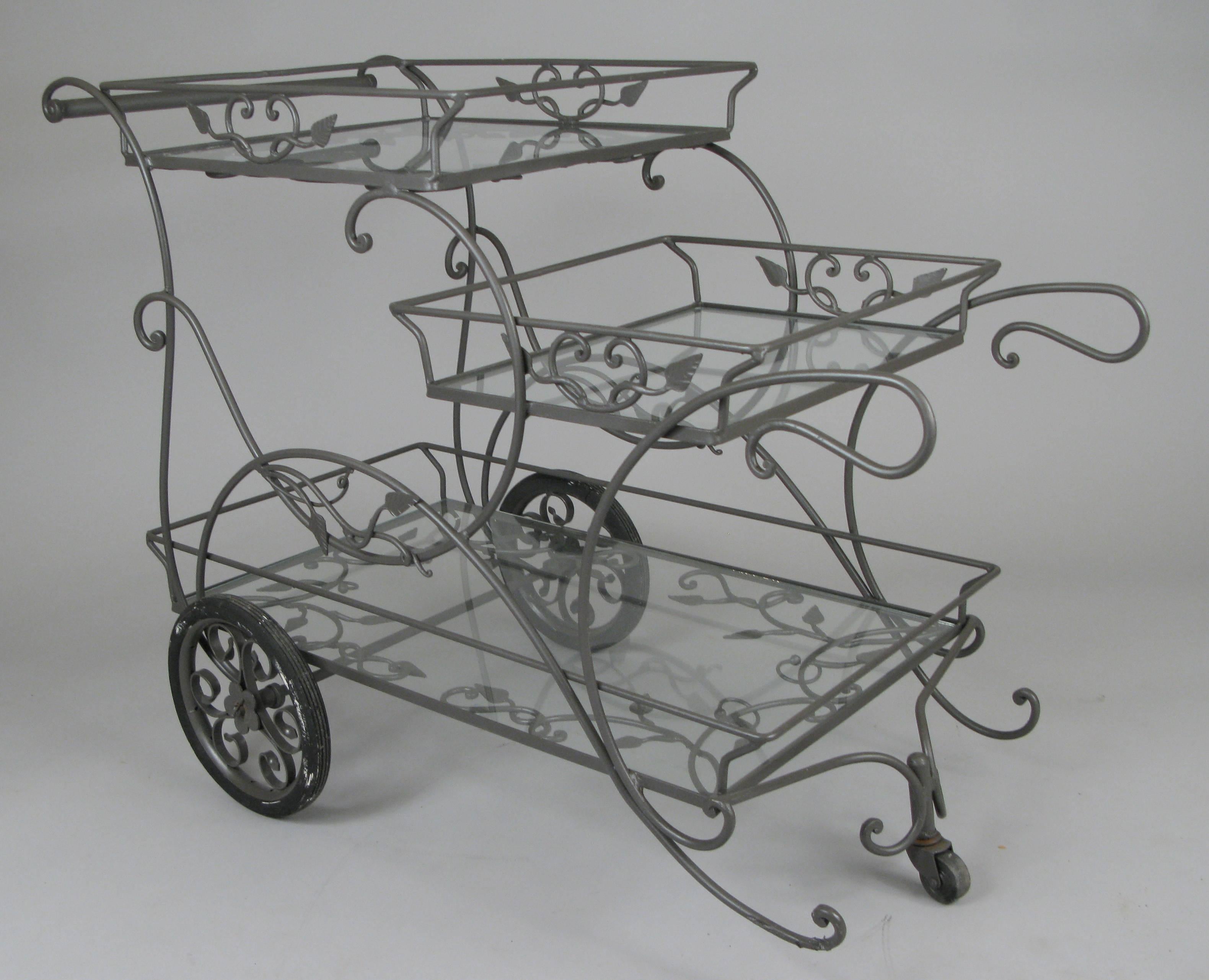 A beautiful vintage 1950s rolling bar cart in wrought iron by Woodard, with three glass shelves for plenty of room for bottles and glassware. The frame decorated with scrolls and ivy. Finished in a greige painted finish.