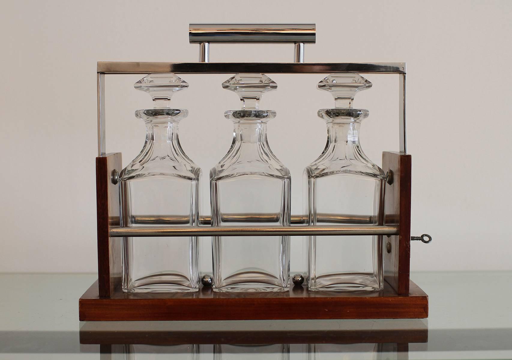 French Art Deco Tantalus decanter set attributed to ADNET, three crystal bottles enclosed in a burl Amboyna and chrome-plated frame.
A lock and a key allow to swing the handle to the back and release the bottles kept in place and secure.
Original