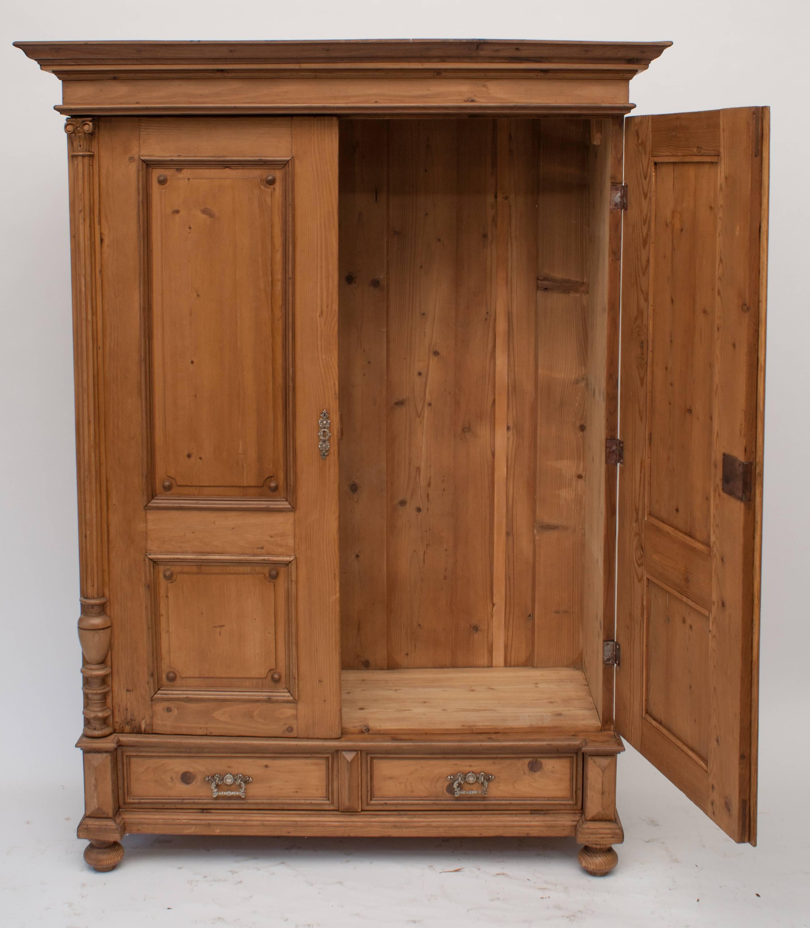 This lovely small pine armoire features a bold hardwood crown and Corinthian split balusters to the side of each wide-swinging door.  Each door has two raised panels with applied hardwood buttons in the corners.  There is a single beautifully hand