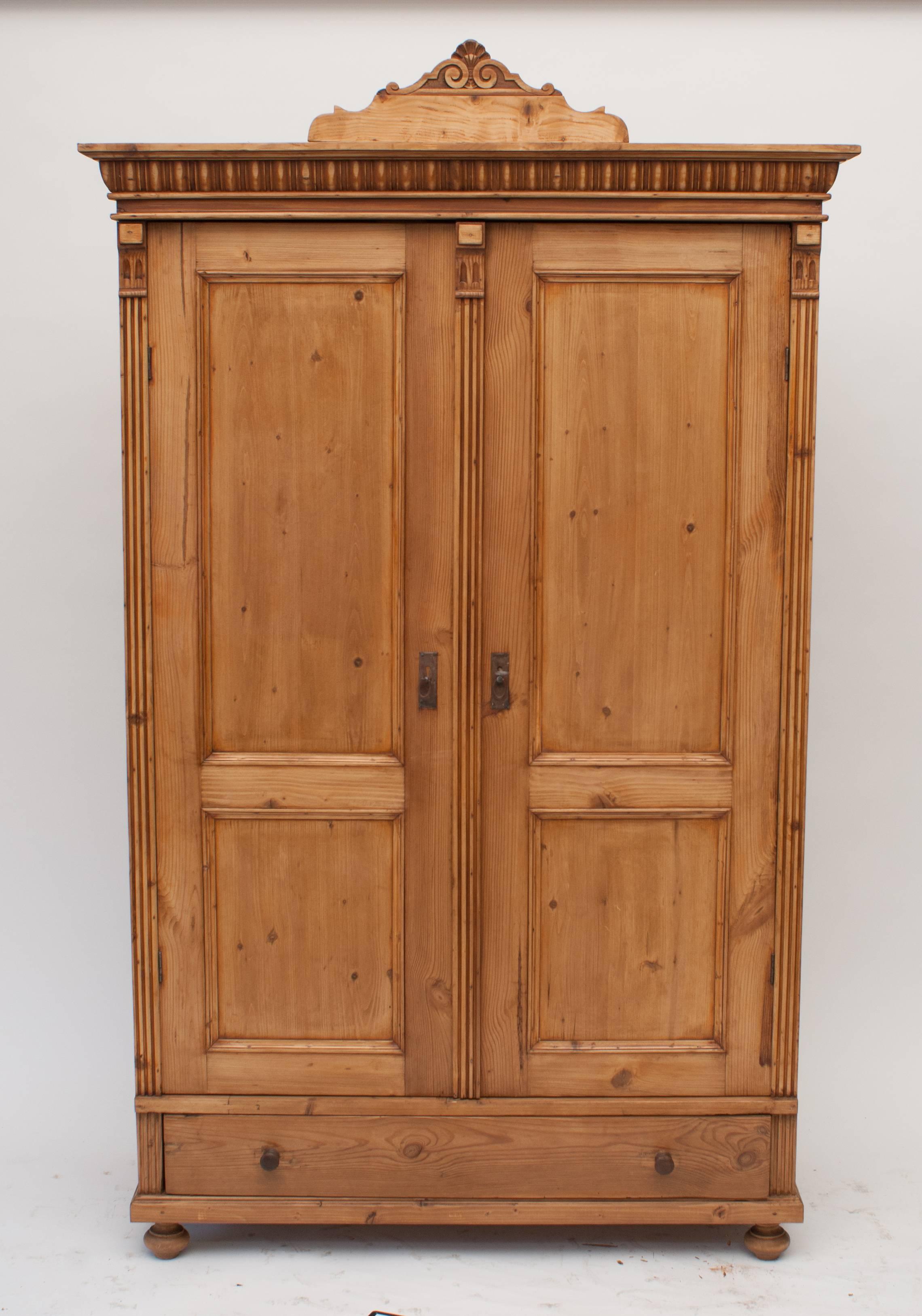 A beautiful small pine two door armoire, each door with two flat panels, flanked and separated by fluted moulding and corbels and opening to a convenient 130 degrees.  Decorative crown moulding is embellished by a removable hand carved crest. 