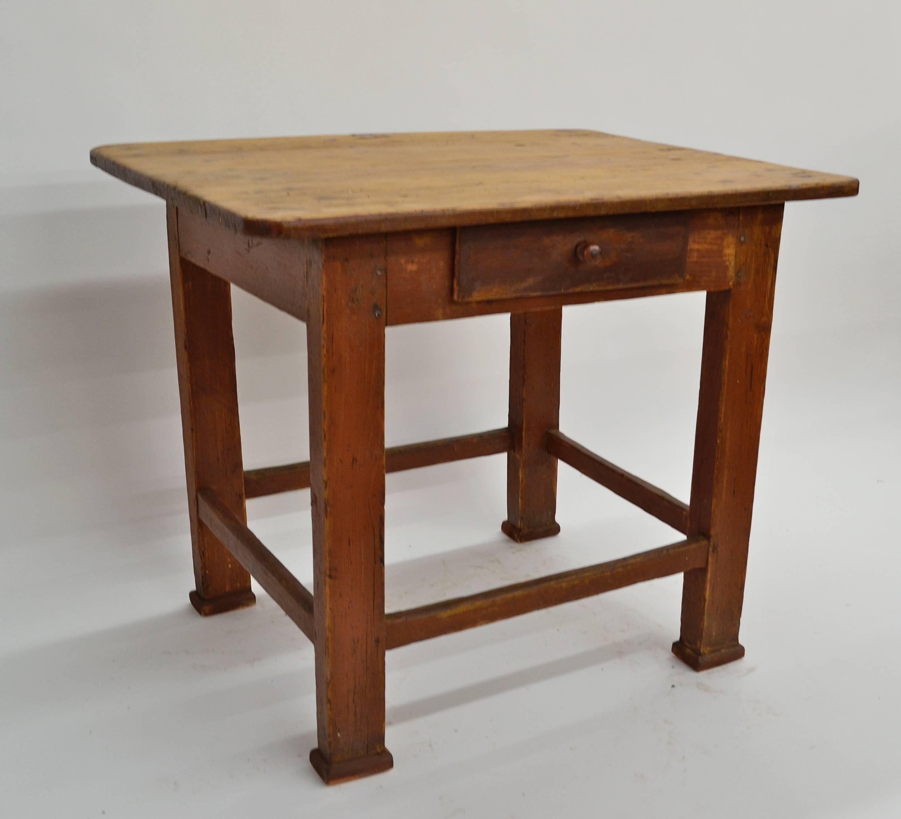 A beautifully worn one drawer pine end table featuring a naturally distressed three board top with smoothly rounded corners. The four straight legs are joined by four worn stretchers. The base is in original oxblood paint rubbed through to bare wood