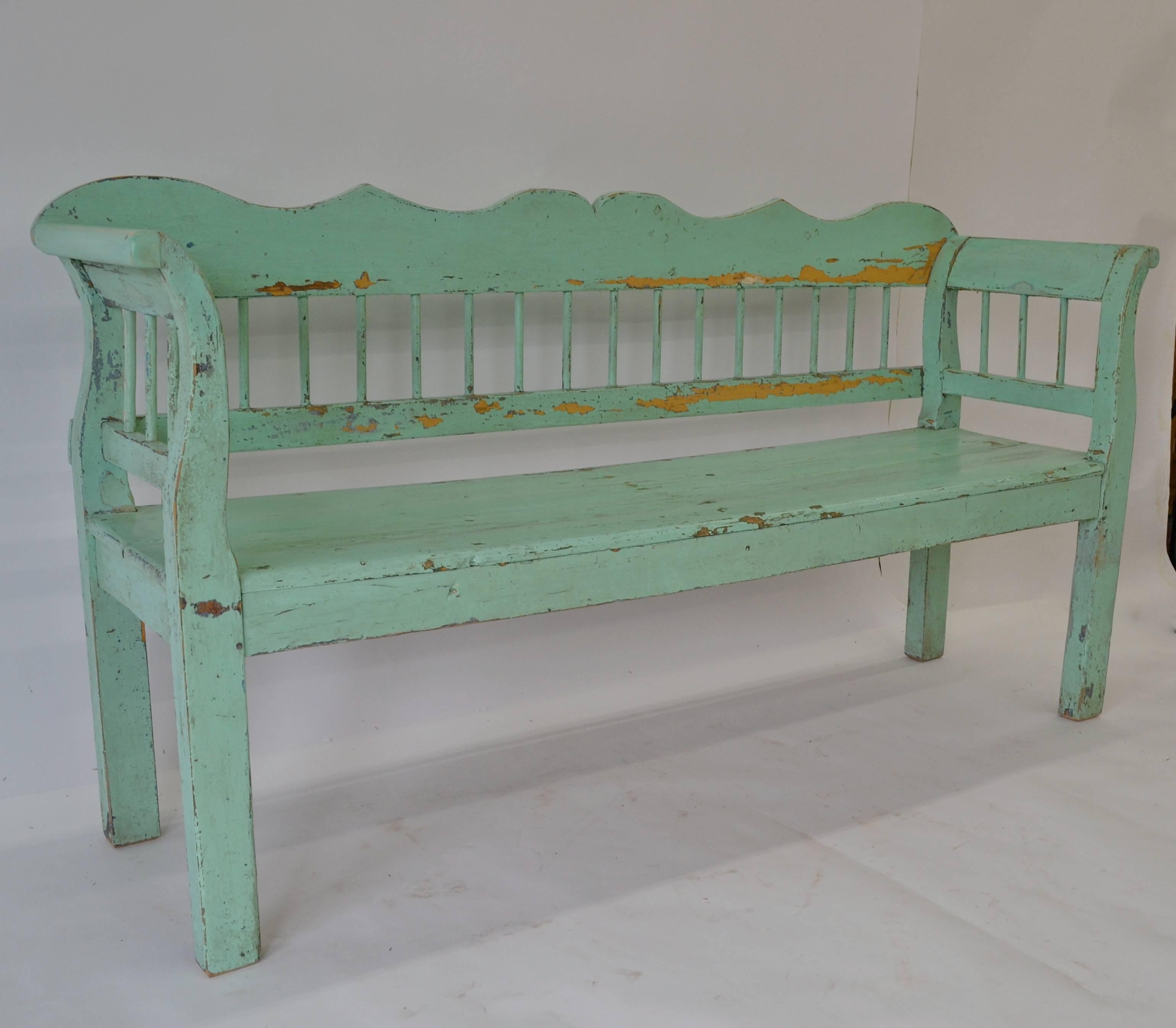 A pine bench or settle with a scalloped top rail and boldly scrolled arms. Plain dowelled spindles join the back and side rails and the generous seat stands on straight legs. In old pale green paint worn and peeled through to earlier light mustard.