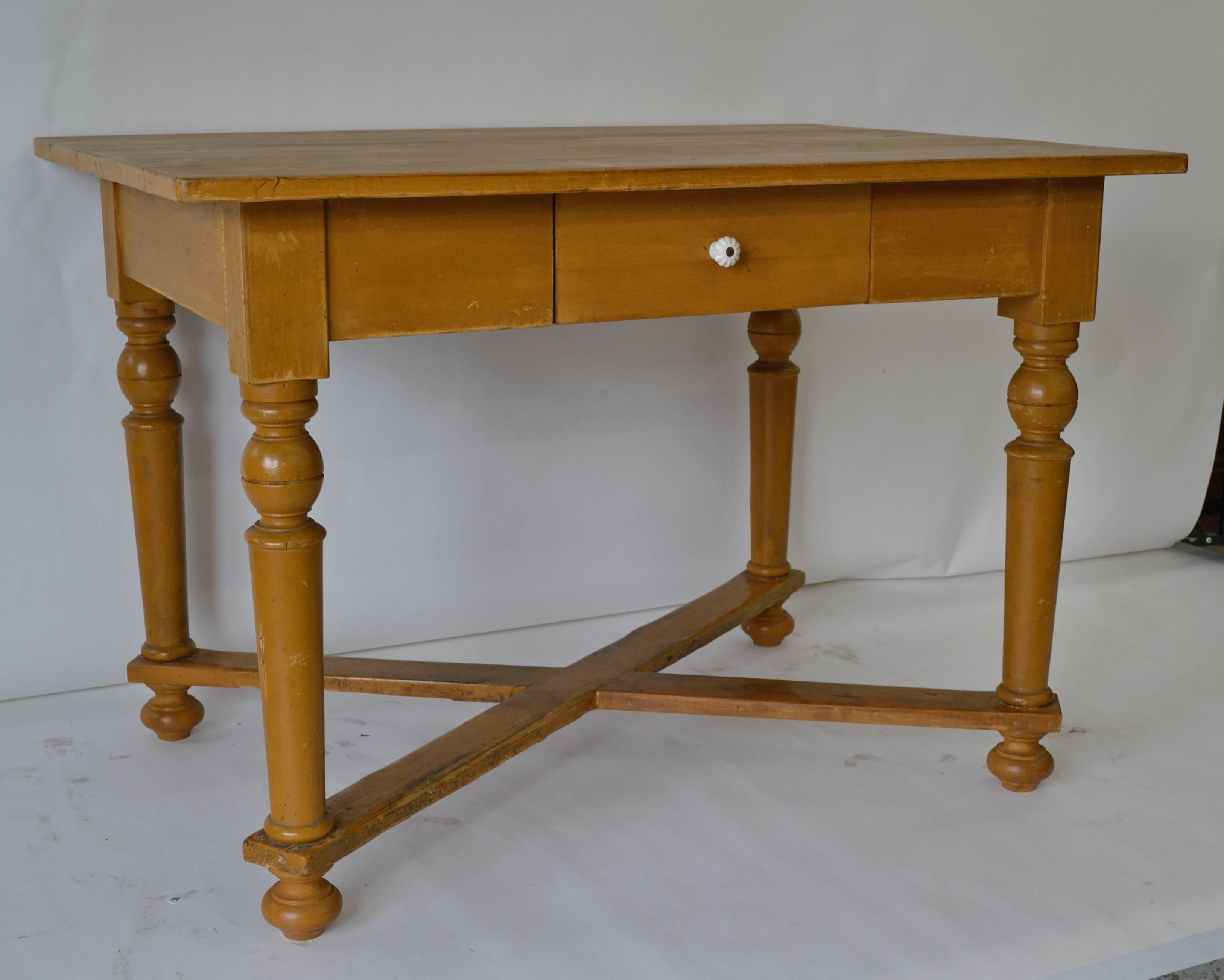 This attractive writing table has four nicely turned legs joined at the foot with a well-worn cross stretcher and a single hand-cut dovetailed drawer in the front apron. The top is in polished pine and the top edge and base in old worn mustard paint.