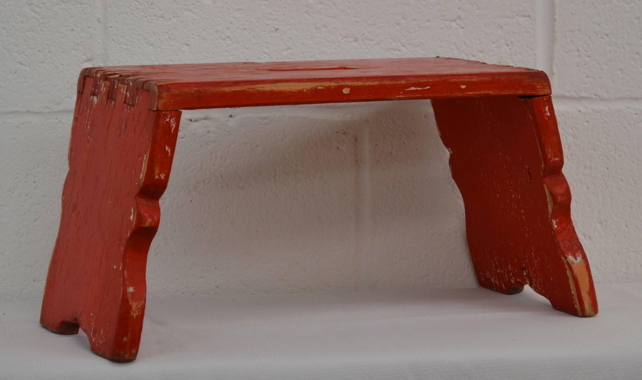 Ein charmanter, schwalbenschwanzförmiger Schemel mit geschwungenen Seiten und ausgeschnittenem Griff.  In altem Rot, das bis zur ursprünglichen weißen Farbe durchgerieben ist.  