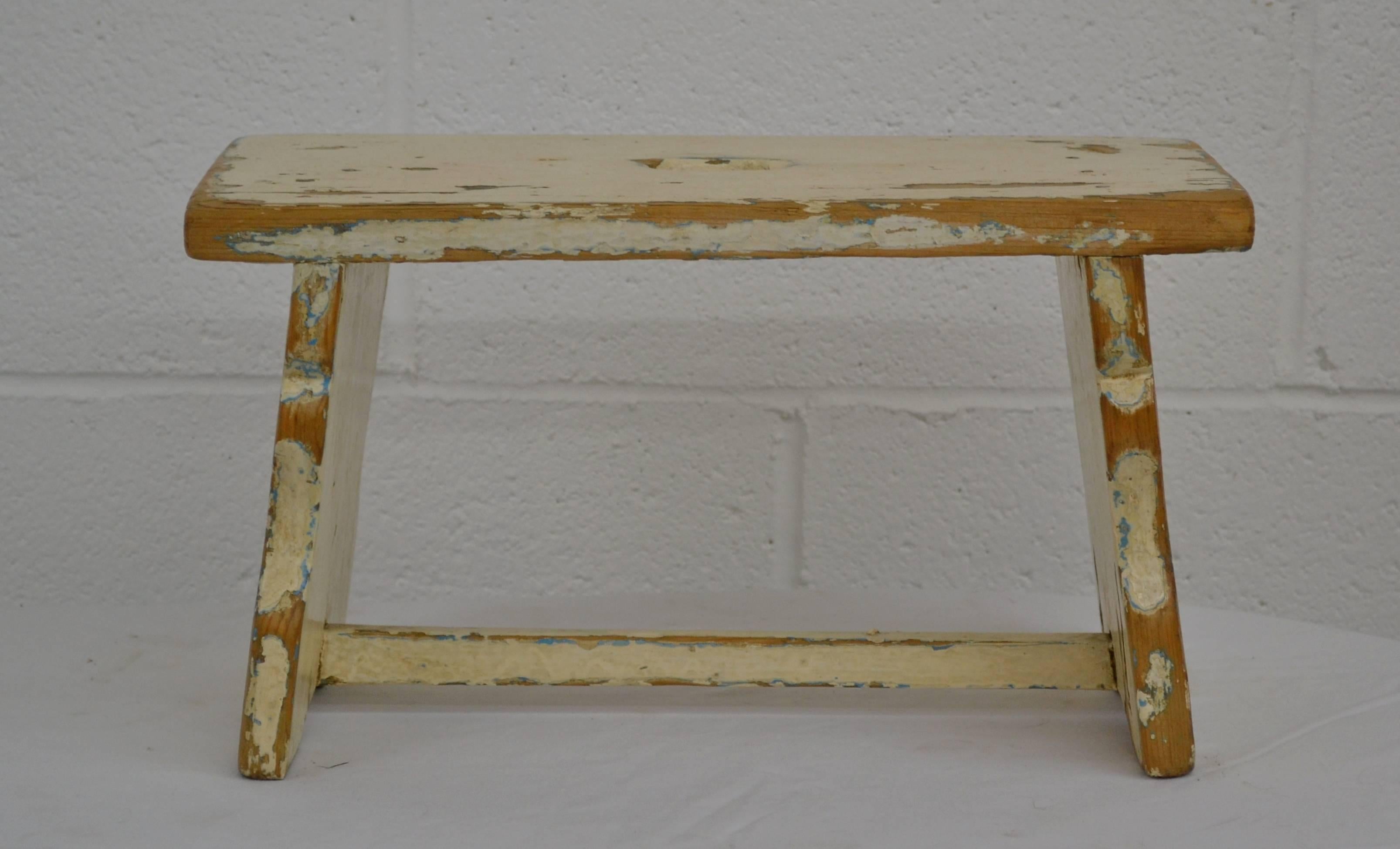 Kleiner Fußschemel aus Kiefernholz mit gezackten Böcken, niedriger Bahre und ausgeschnittenem Handgriff.  In alter cremefarbener Farbe, die bis zum Holz durchgerieben ist.  