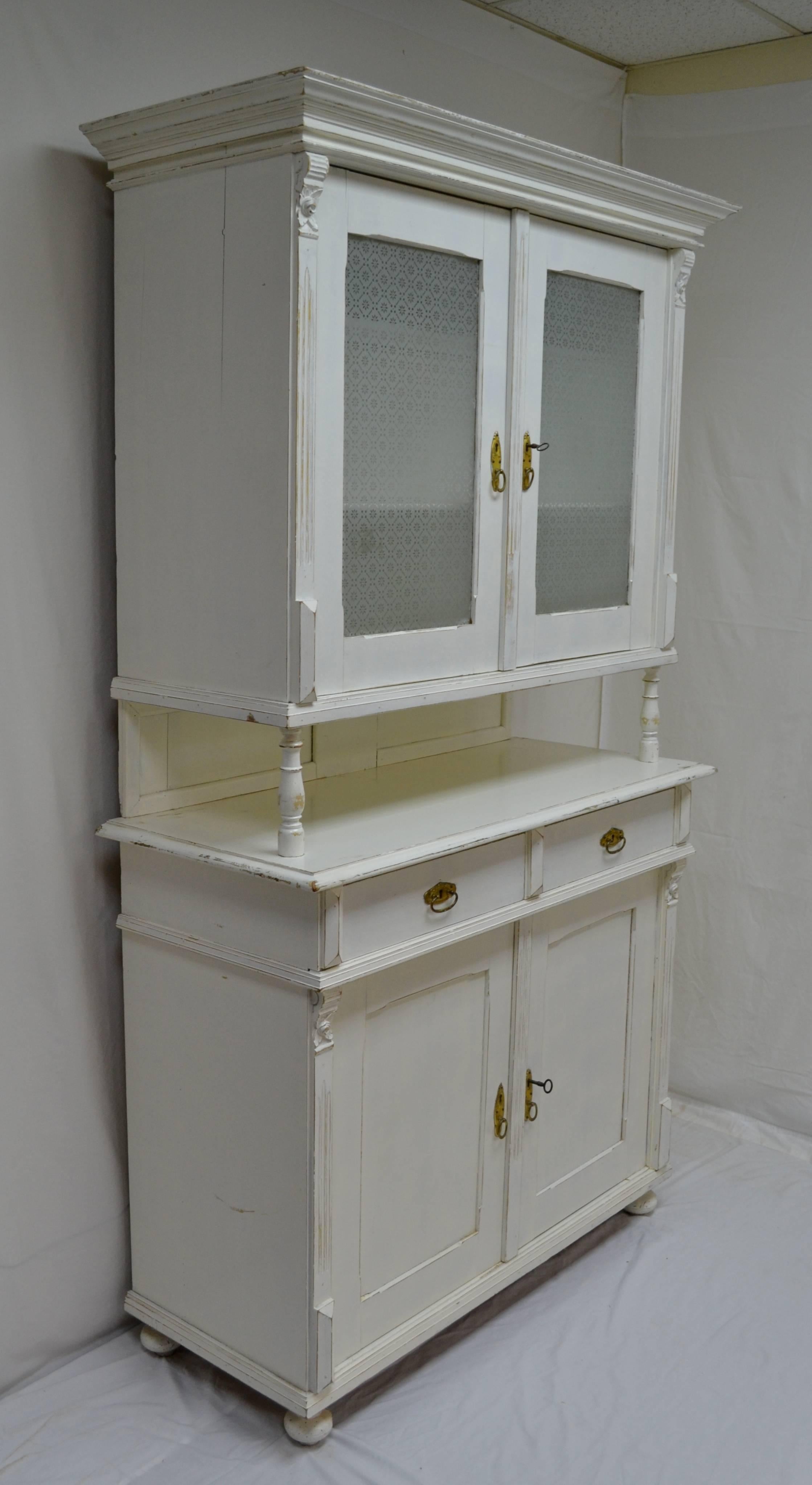 A Classic European buffet or glazed dresser featuring original acid etched glass to the upper section, two hand-cut dovetailed drawers and paneled doors to the lower. Fluting and carved rosebud corbels to the front sides. Later distressed white