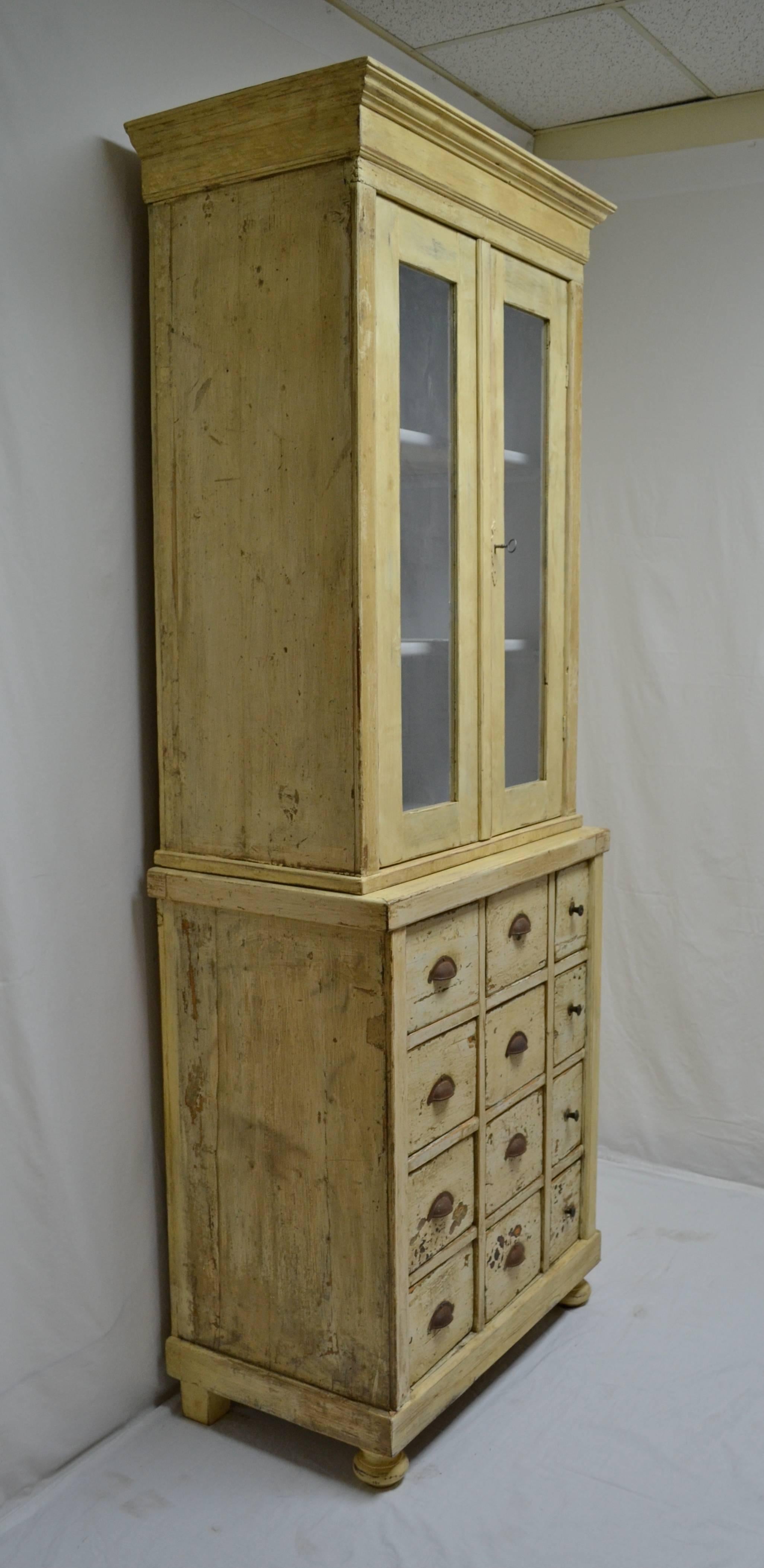 This is an unusually narrow pine apothecary cupboard, built in two pieces. The upper section has two glass doors which open wide to a spacious two shelf interior, fairly recently painted in pale blue. The base has twelve deep original hand-cut
