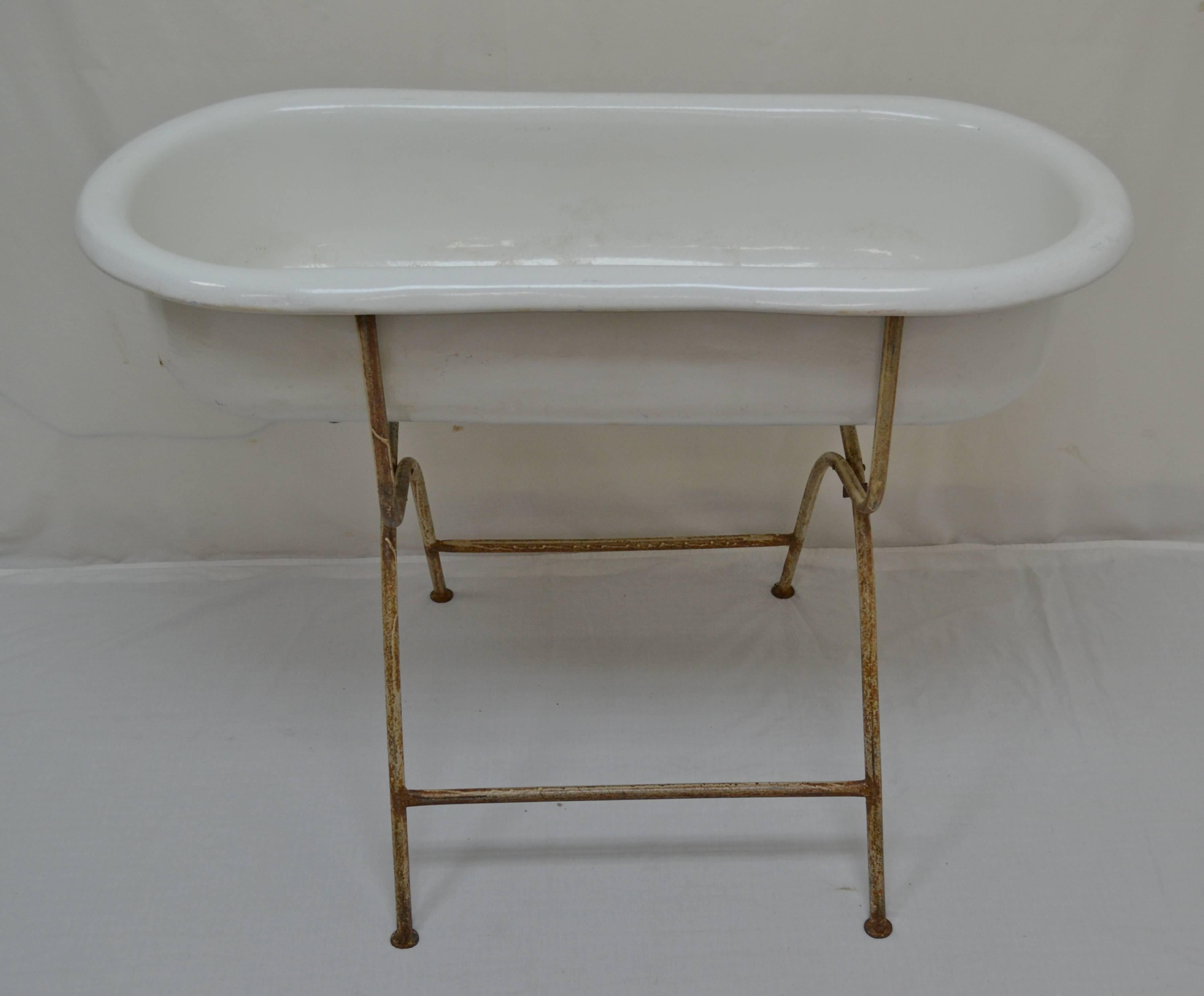 An attractive vintage enameled baby bath by “Zim” of Hungary, on its original tubular steel folding stand. There are some small scratches and repairs to the enamel. The stand is in old worn and rusted cream paint. The drain tap is missing but could
