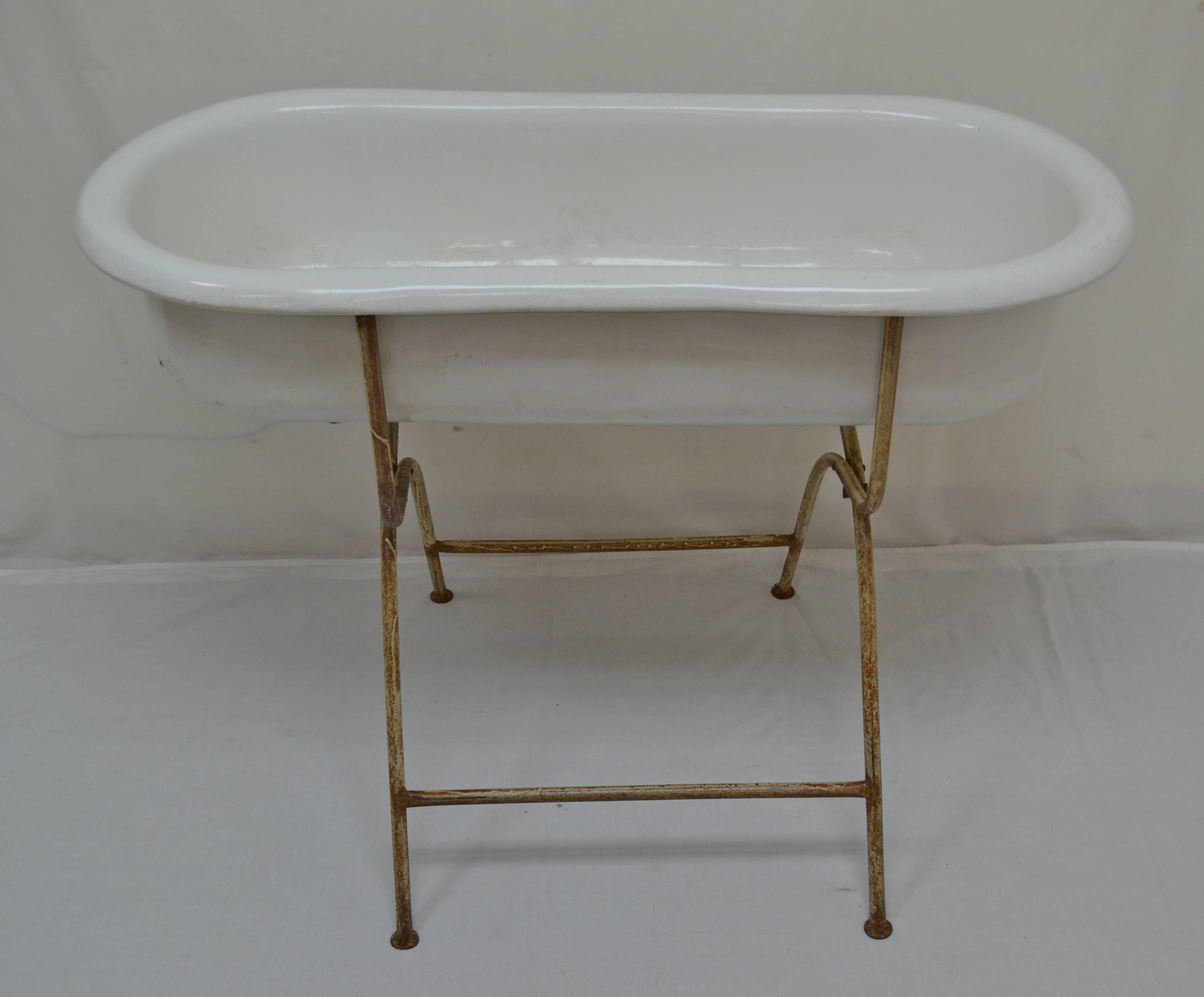 An attractive vintage enameled baby bath by “Zim” of Hungary, on its original tubular steel folding stand. There are some small scratches and repairs to the enamel. The stand is in old worn and rusted cream paint. The drain tap is missing but could
