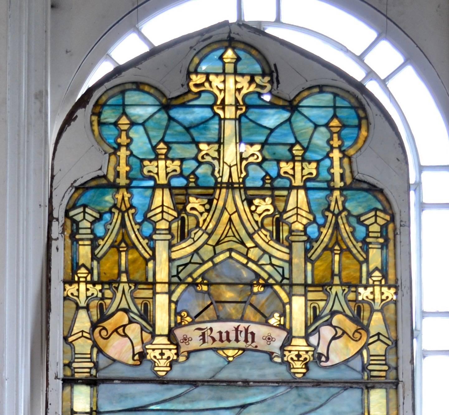 Eine massive gotische, farbenfrohe Glasmalerei, die die biblische Ruth mit einer Weizengarbe in einem Weizenfeld an einem Bach zeigt.   Das Bild wird von Säulen umrahmt, die vor einem blauen Himmel einen kunstvollen Turmkranz tragen.  Ausgiebige
