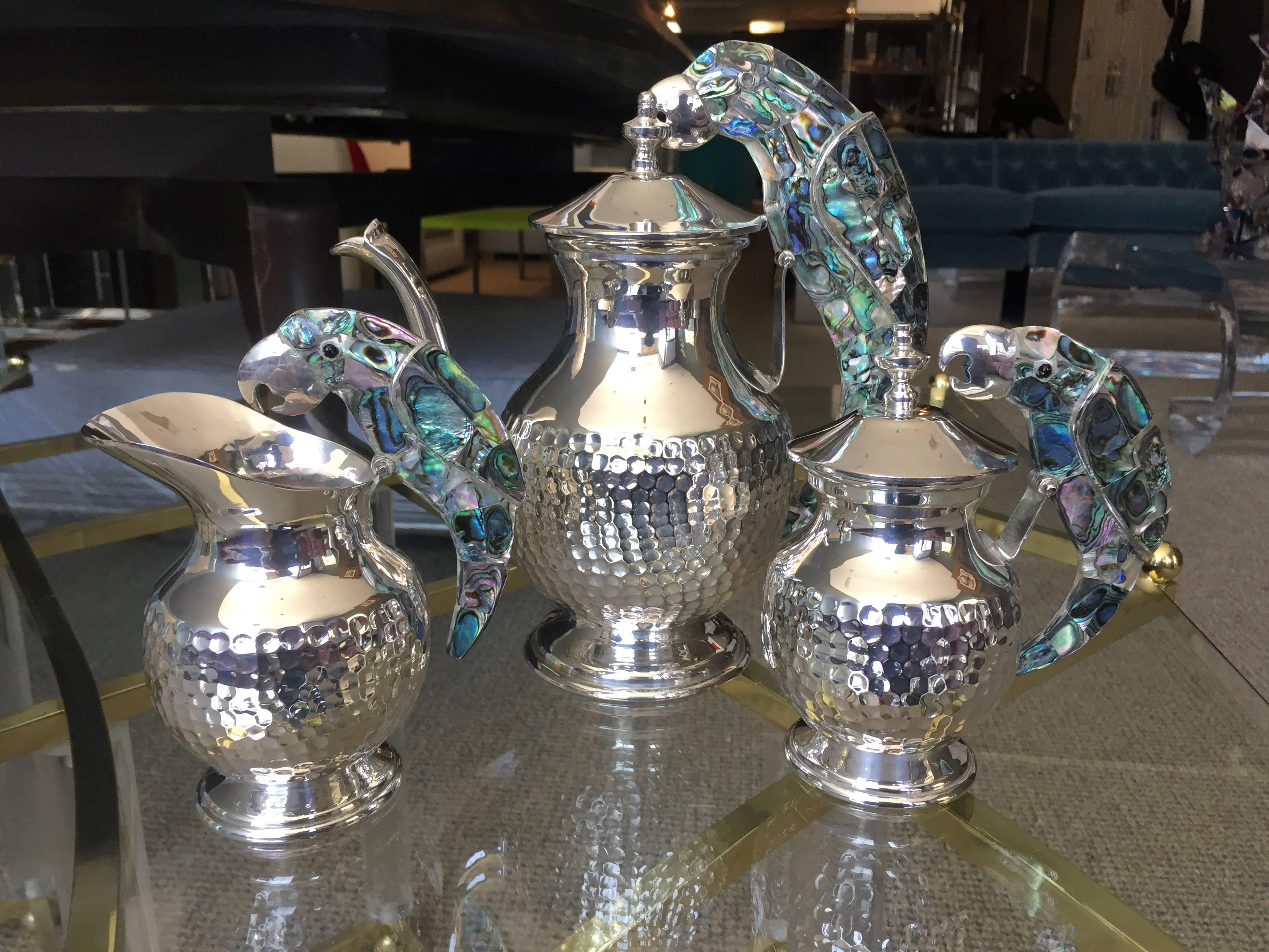 Beautiful and rare tea service set in silver and abalone shell by the renowned silversmiths Los Castillo from Taxco Mexico.
This beautiful set consist of a tea pot a creamer and a sugar pitcher, every piece is beautiful decorated with a parrot