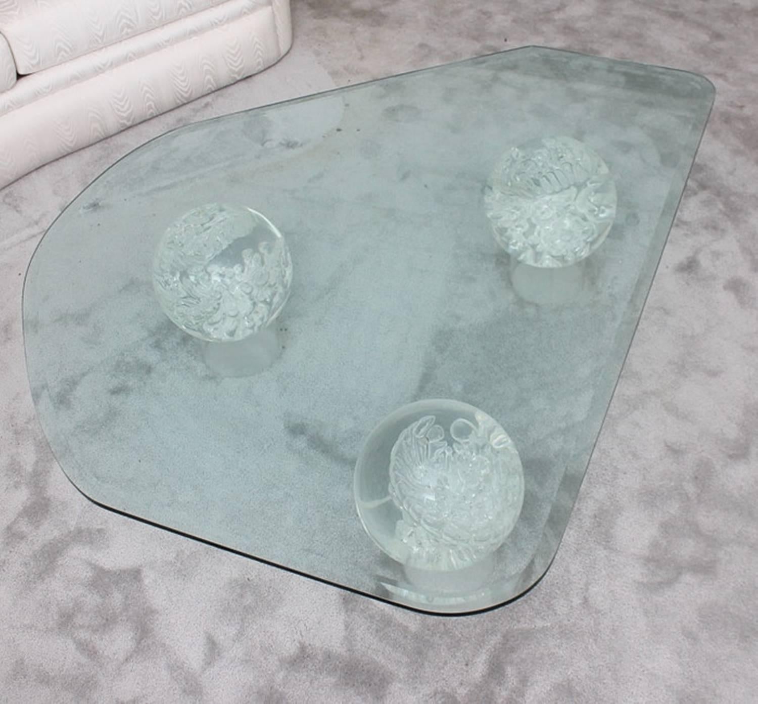 A Mid-Century Modern glass coffee table. This piece features an irregular shaped top, with a beveled edge. The top is elevated on three glass spheres, with bubbles and swirls visible within. Each sphere rests on a clear acrylic cylindrical base. The