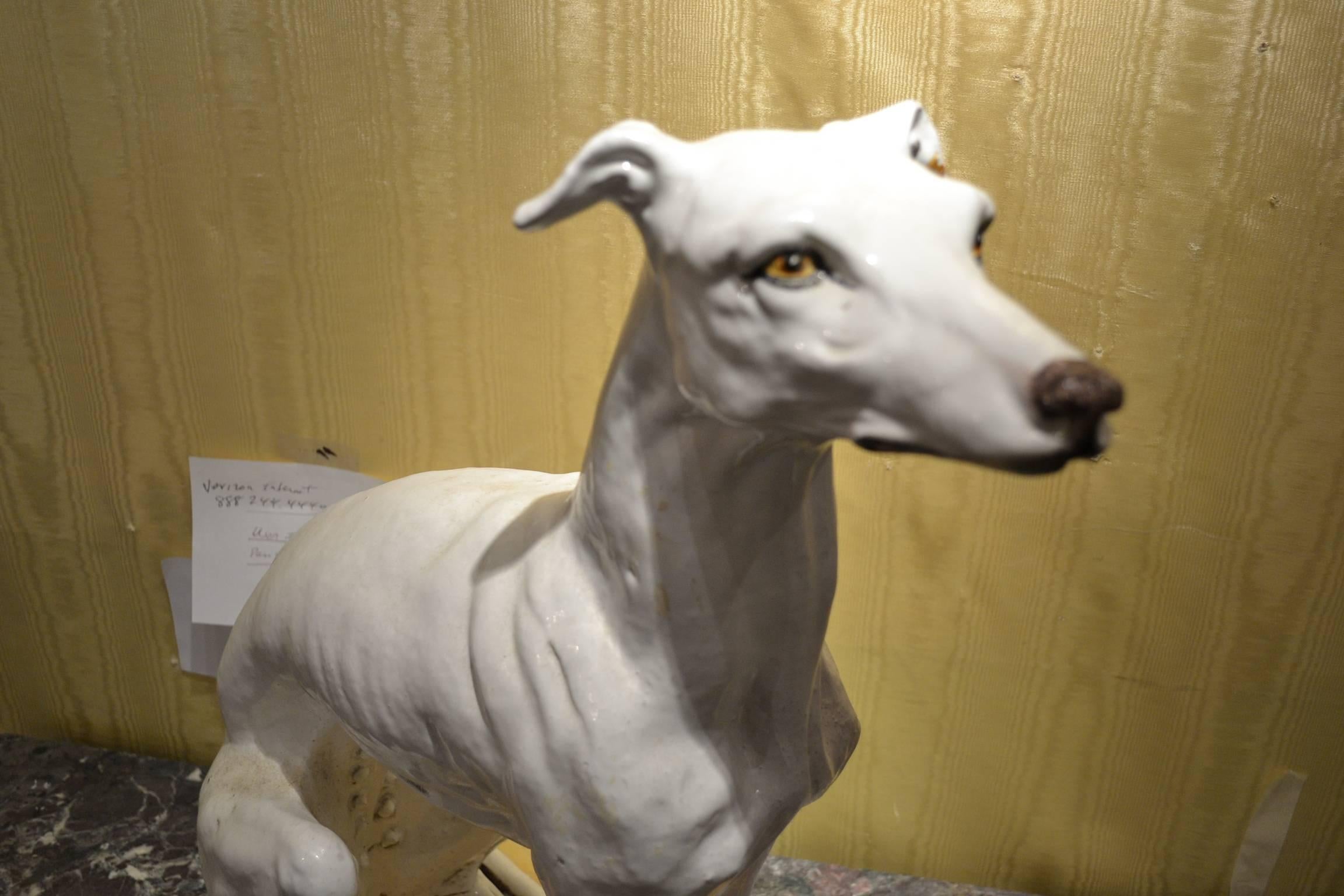 Terracotta seated figure of a greyhound dog on pillow with fringes on each corner with various cracks.