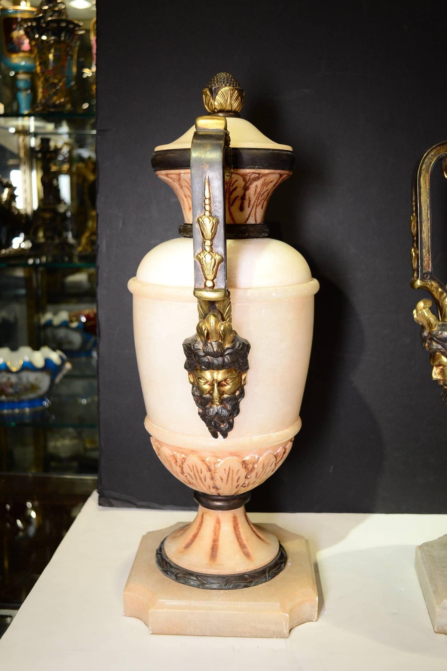 Neoclassical pair of carved alabaster and gilt bronze painted urns with mask face handles on square form bases.