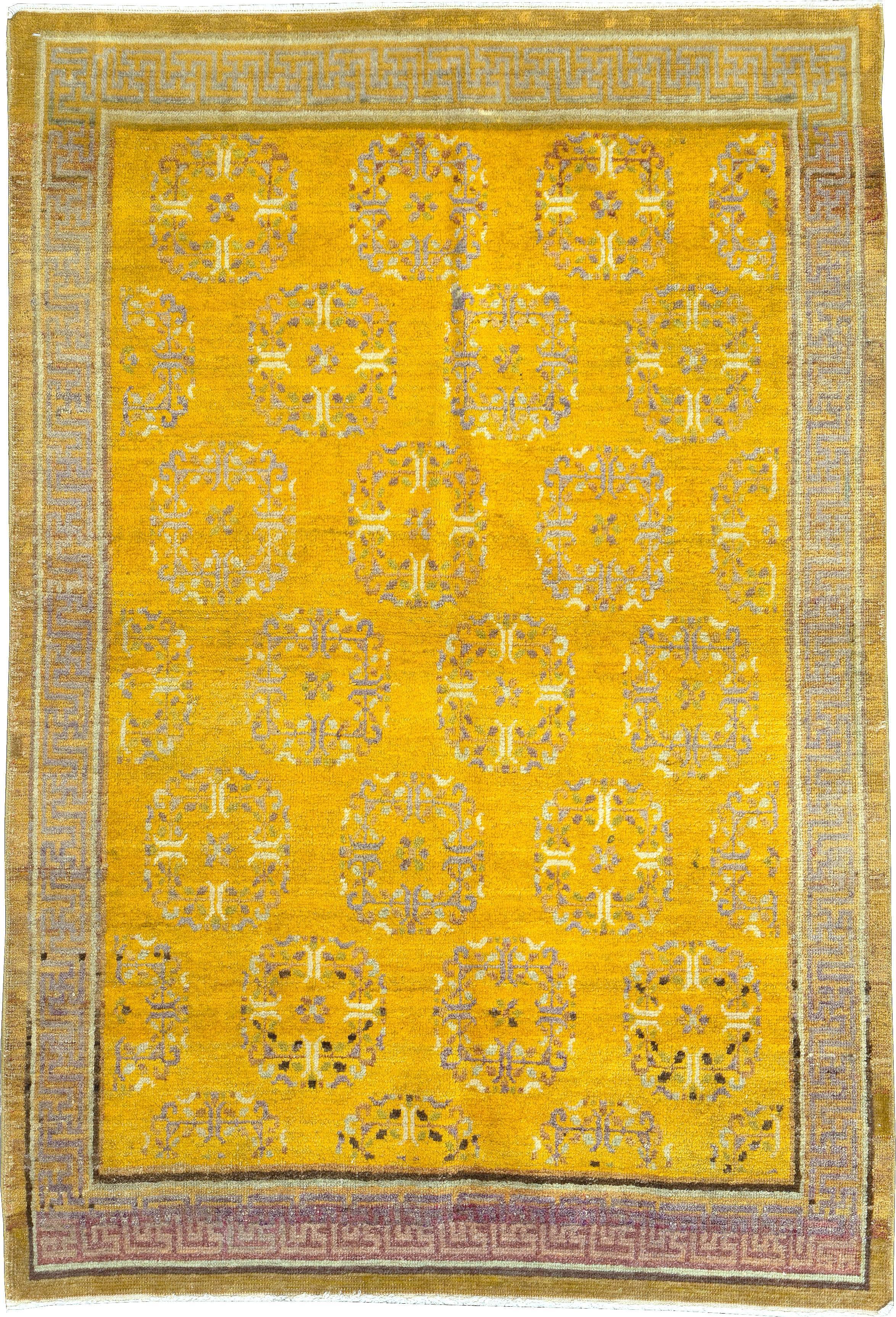 An antique East Turkestan Khotan carpet from the early 20th century. The saturated goldenrod ground displays offset rows of floral wreath roundels, set off by a slate and pomegranate colored swastika fret border. The gold outer band is plain, and
