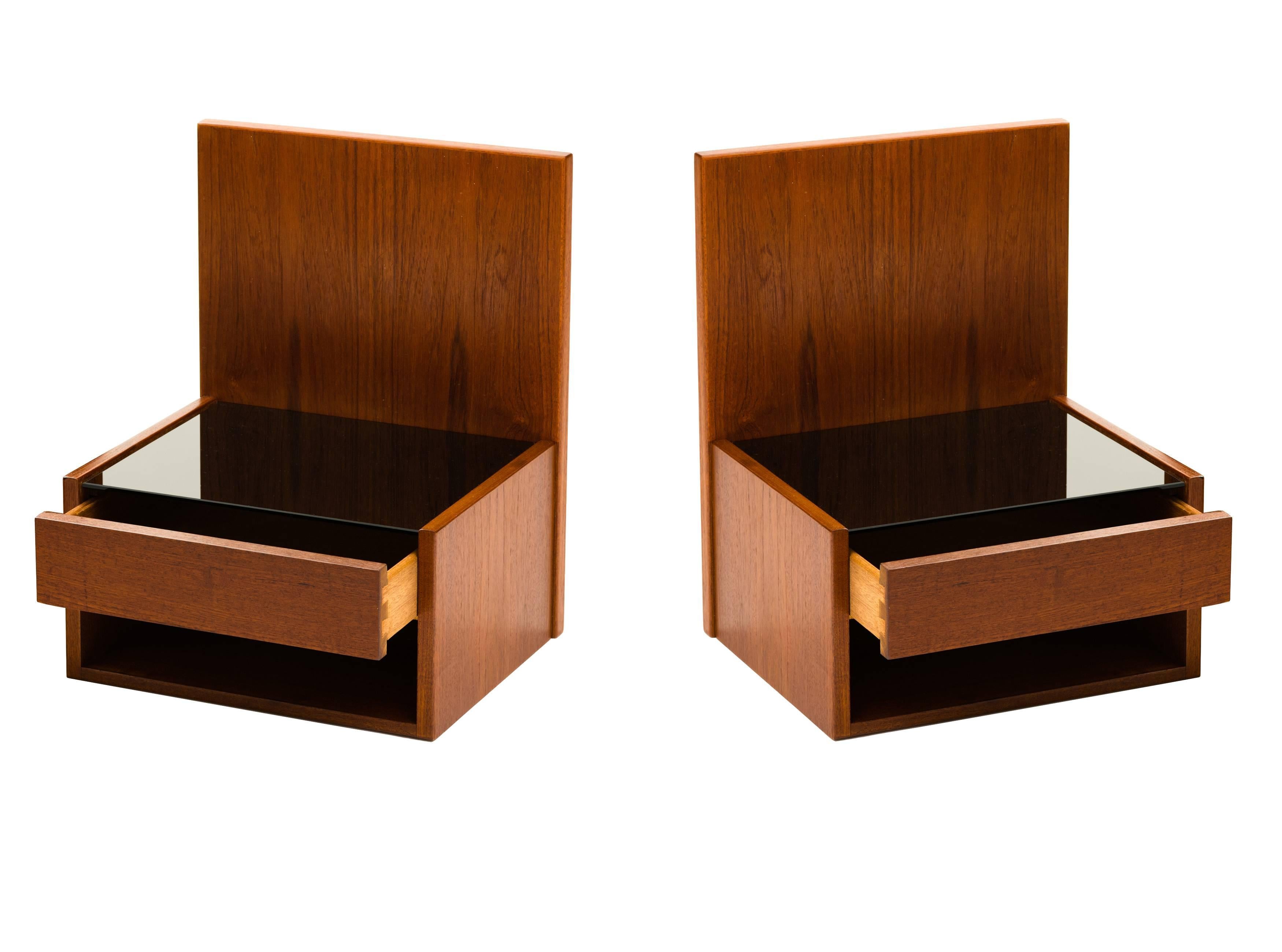 A pair of floating night stands or shelves in teak, each with a single drawer, open storage compartment beneath and a smoked glass top surface, designed by Hans Wegner for Getama. These may be mounted either to the wall or to the sides of your
