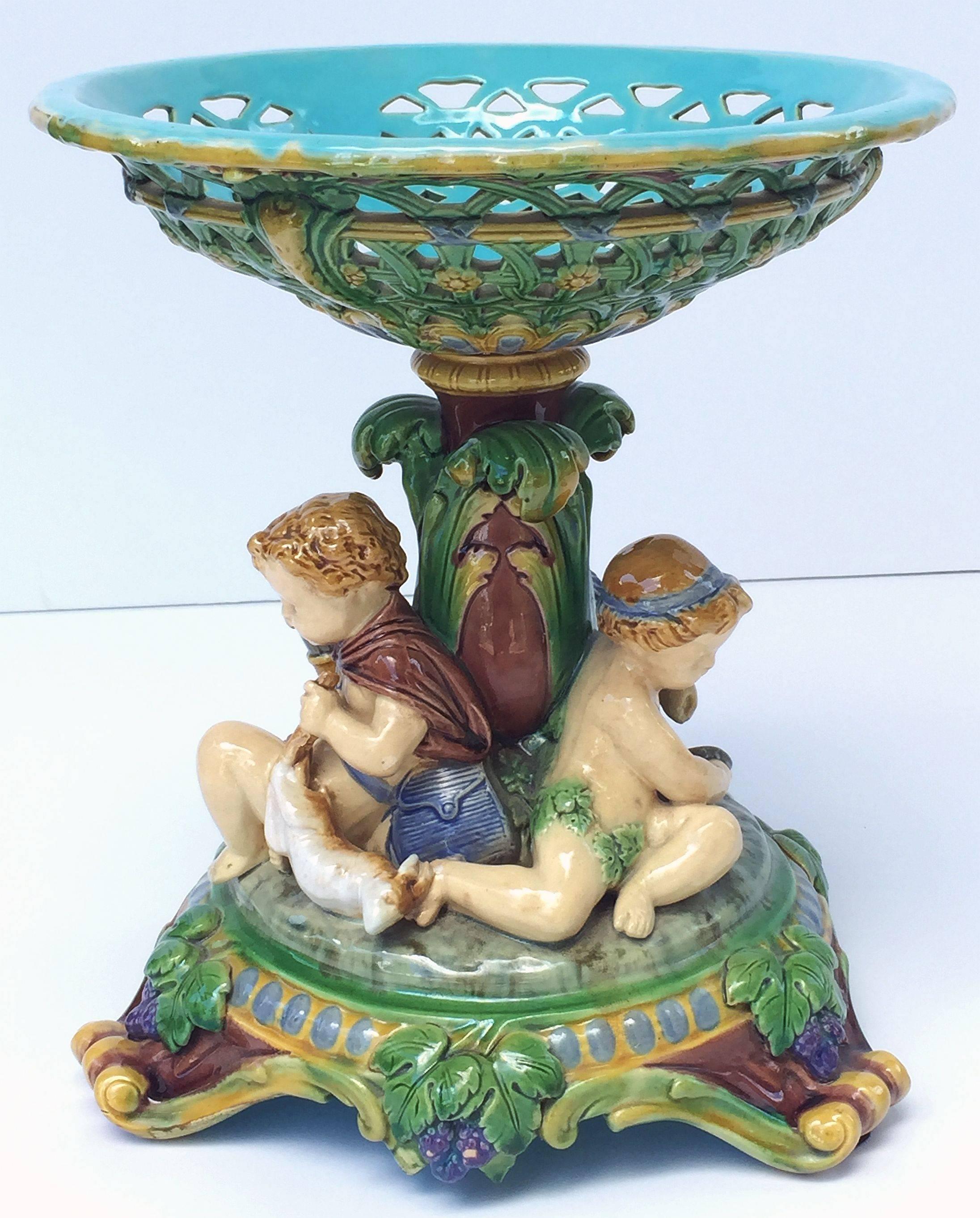 A fine Majolica centerpiece or compote (or comport) table center by the celebrated English pottery firm, Minton.

It features a concave top over a pedestal base with three seated cherub figures in the classical style. The underside of the top with