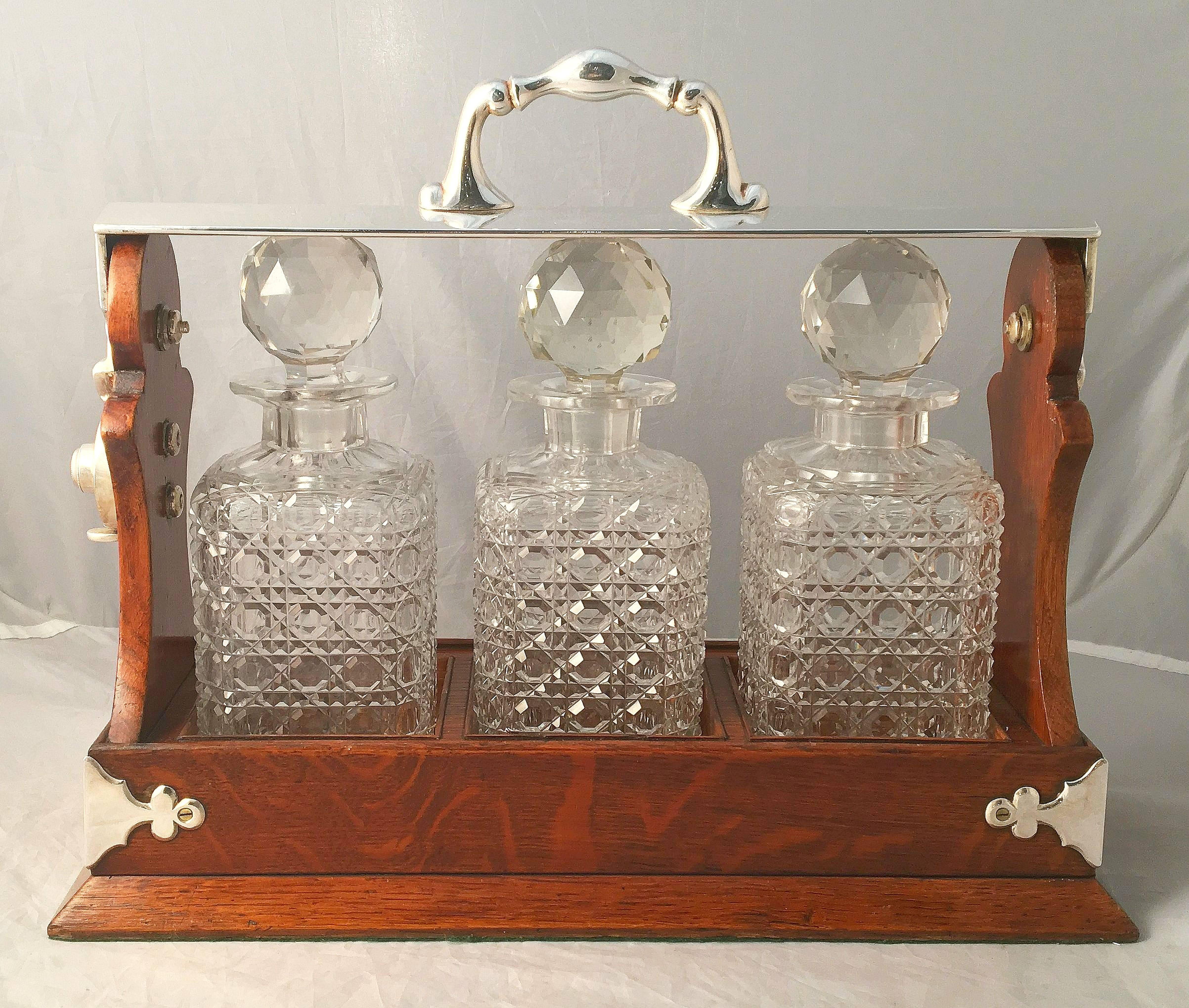 A fine English tantalus or decanter of oak, for whiskey and spirits, from the Edwardian era. 
Featuring Fine plate silver corner brackets, lock mechanism and sturdy carry handle. 
The frame of oak with three fitted cut-crystal decanters with