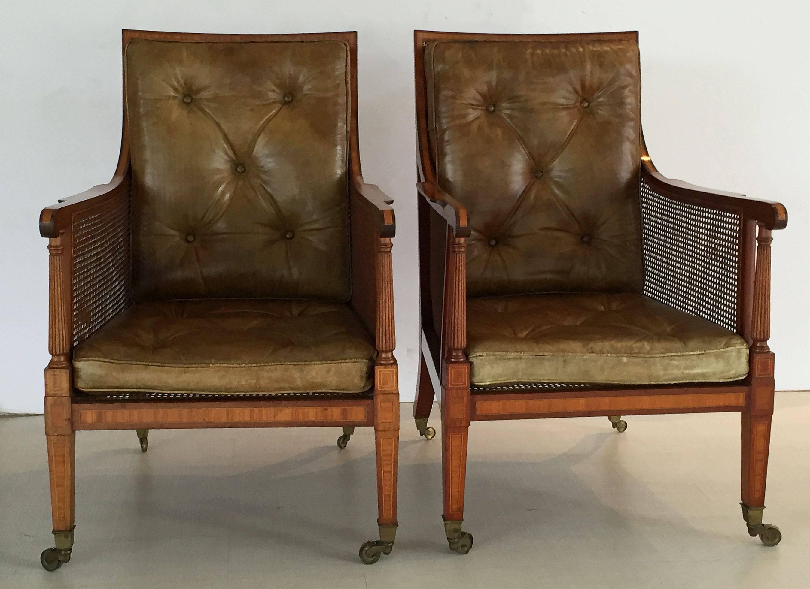 A handsome pair of fine English caned library bergere armchairs from the Edwardian era, each chair featuring inlaid mahogany frames with caned back, seat, and sides. With turned column arm supports and removable tufted leather cushions for the seat