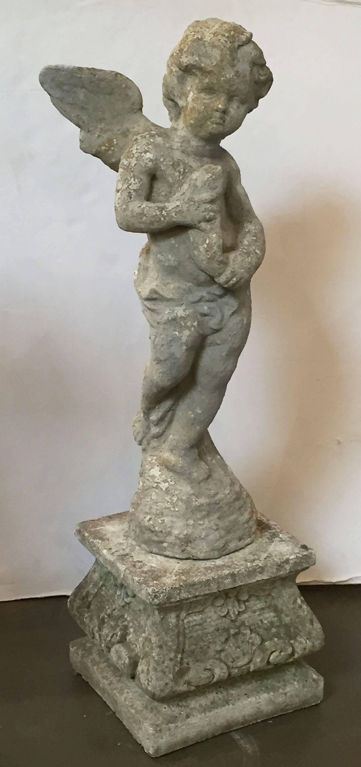 A fine English garden ornamental statue of weathered composition stone, featuring a winged angel holding a fish, standing on a separate square plinth. The plinth base with a relief design of scrolls and seashells.

Perfect for a garden room or