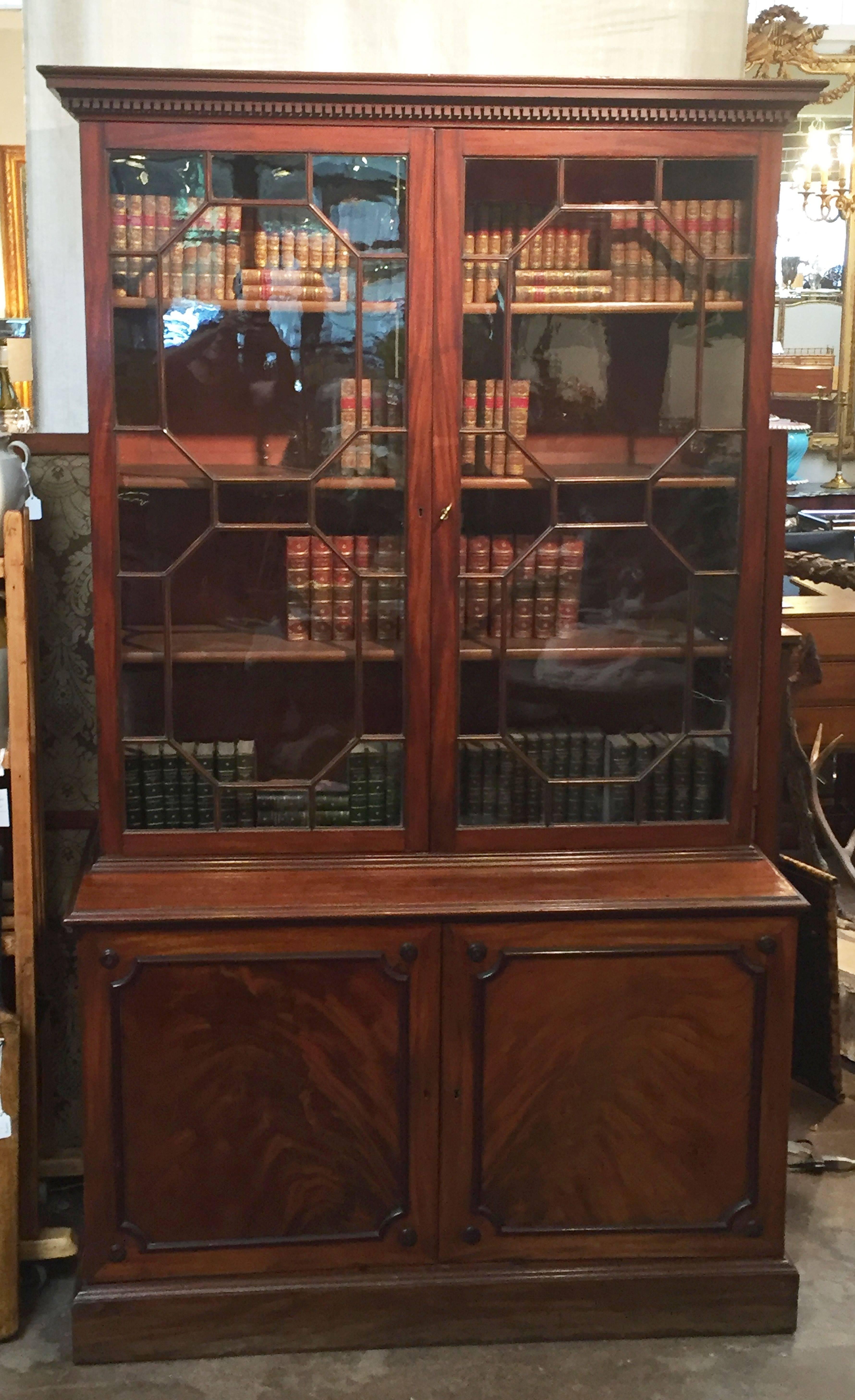 A fine large English bookcase of mahogany, featuring an upper tier with cornice and dentile moulding, two astragal glazed doors enclosing three adjustable bookshelves and the lower cabinet with two-paneled doors of flame-cut mahogany, fitted with