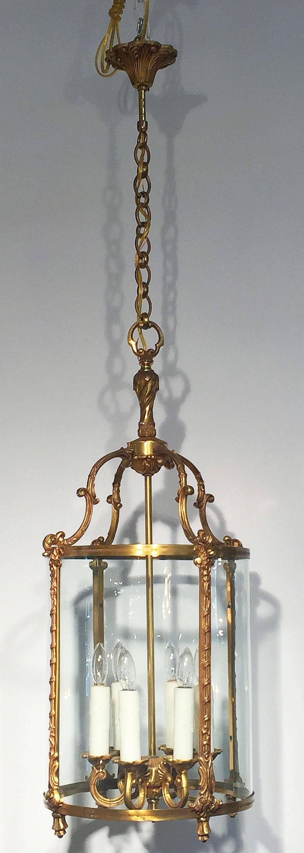 A large French six-light hanging fixture or lantern of gilt metal and glass, featuring a cylindrical body with scroll-work top and six candle lights with gilt metal bobeches and curved glass. Decorative flourishes to top and base. 

Measures:  H