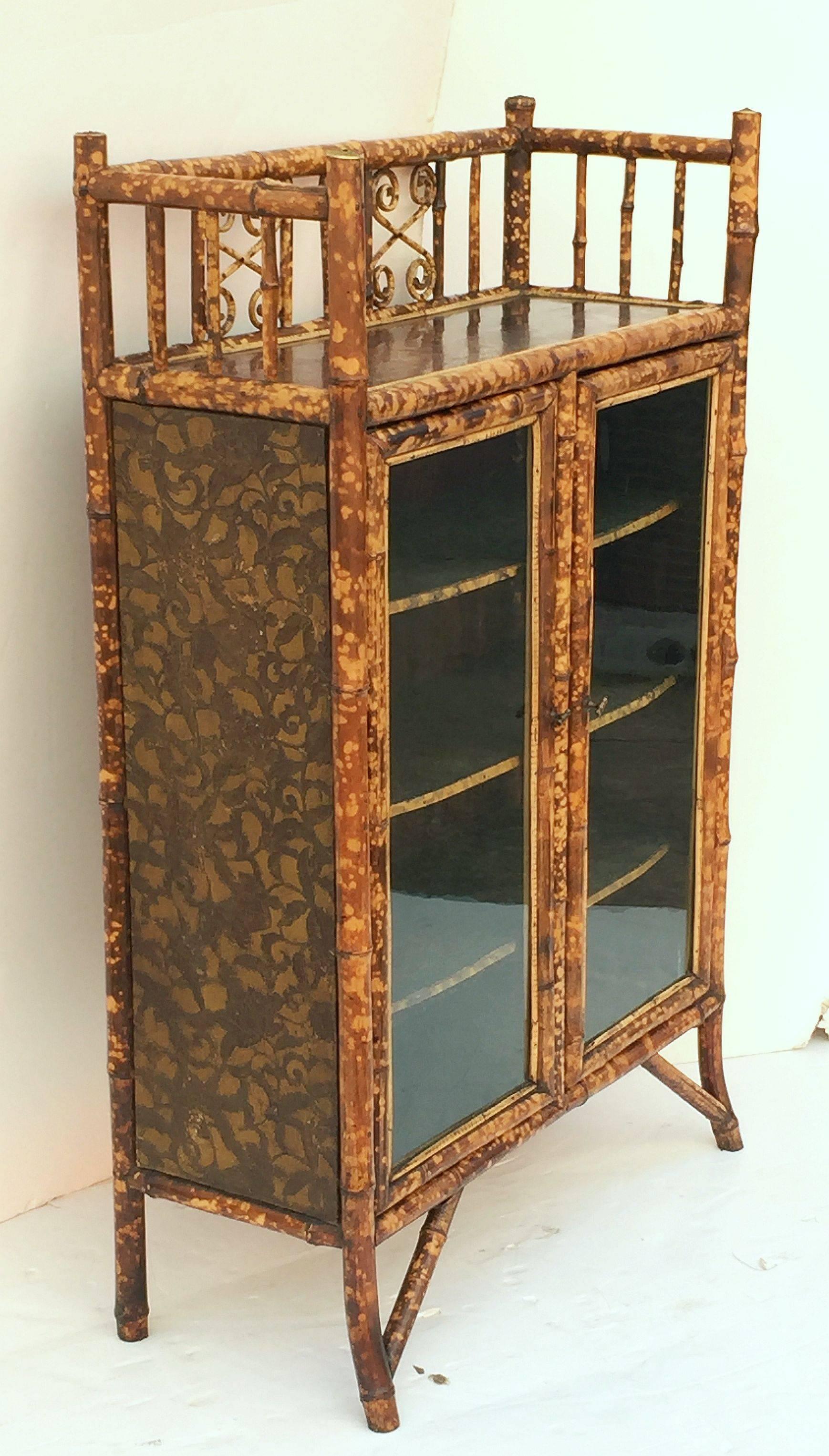 A handsome English bamboo bookcase featuring a tall gallery top of bamboo with bent bamboo accents and lacquered panel, over two framed-glass doors with brass hardware, opening to an interior of three bamboo-edged shelves, each lined with pressed