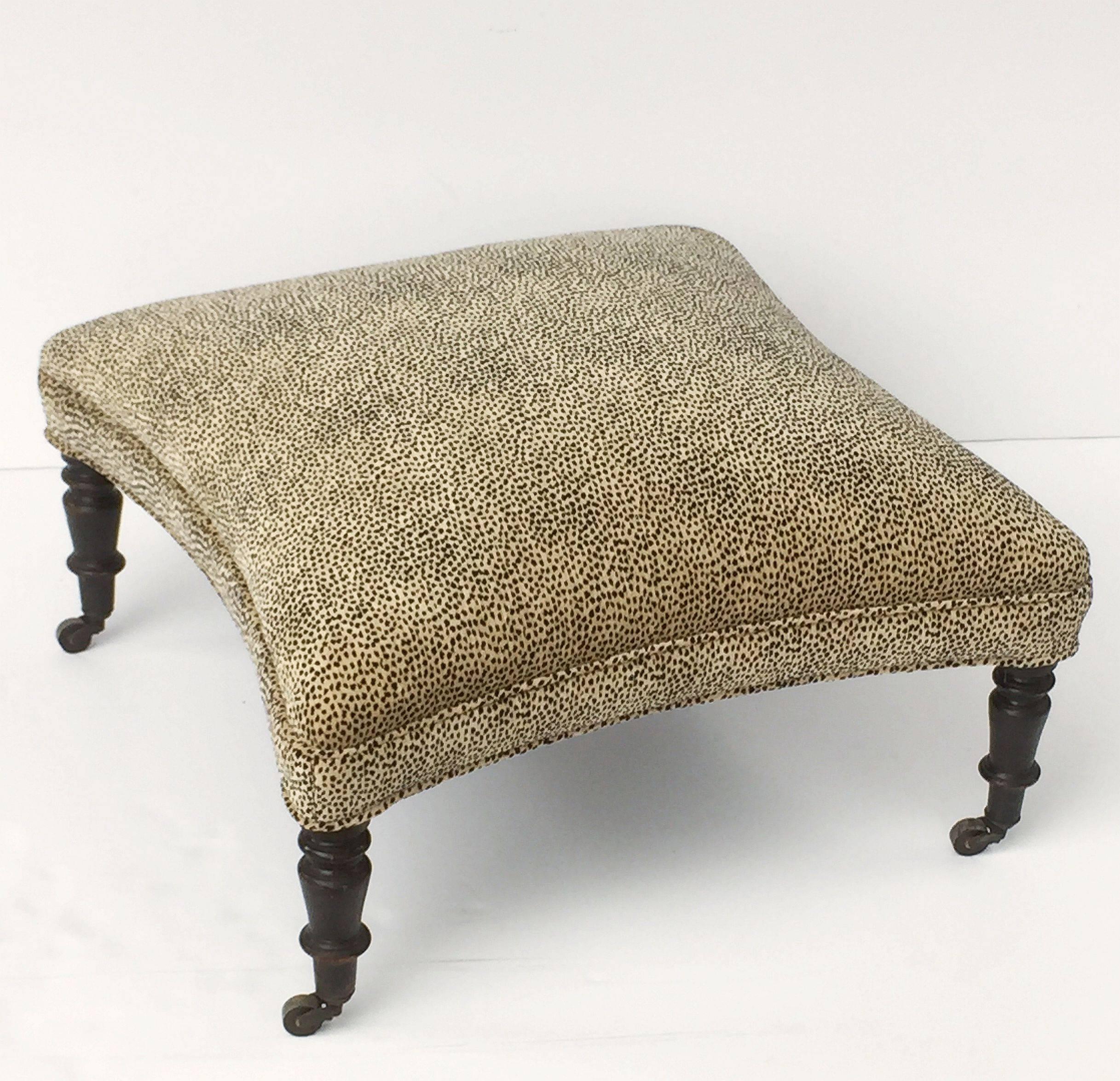 A handsome English upholstered ottoman (or stool) featuring a leopard print fabric on a wood frame with four turned ebonized wood legs, each with rolling brass casters.