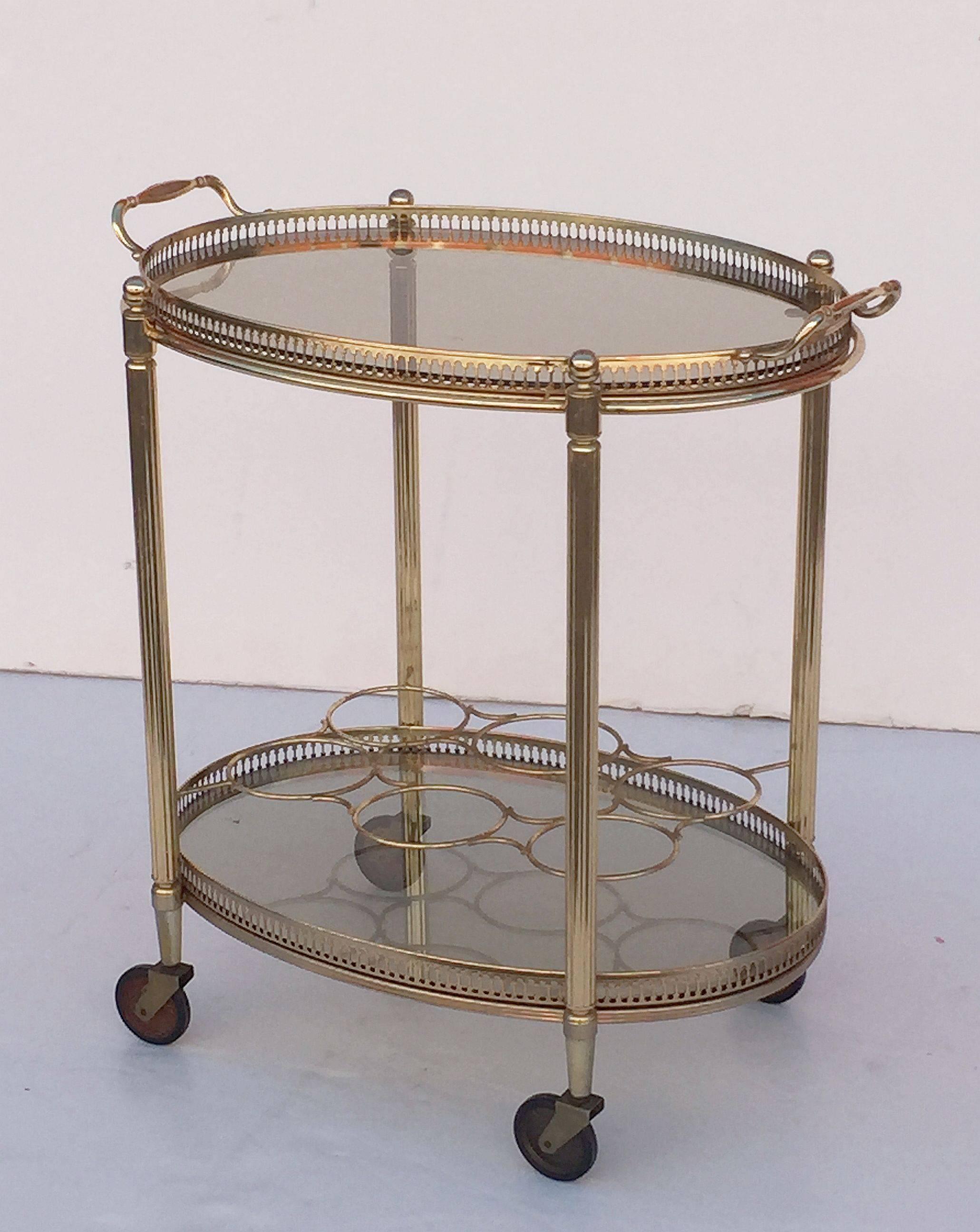 A vintage French oval bar cart table or serving trolley in brass and featuring two smoked glass tiers, each with pierced galleries, with bottle rack for six bottles on second tier, on rolling caster wheels. 

The top tier is removable for use as a