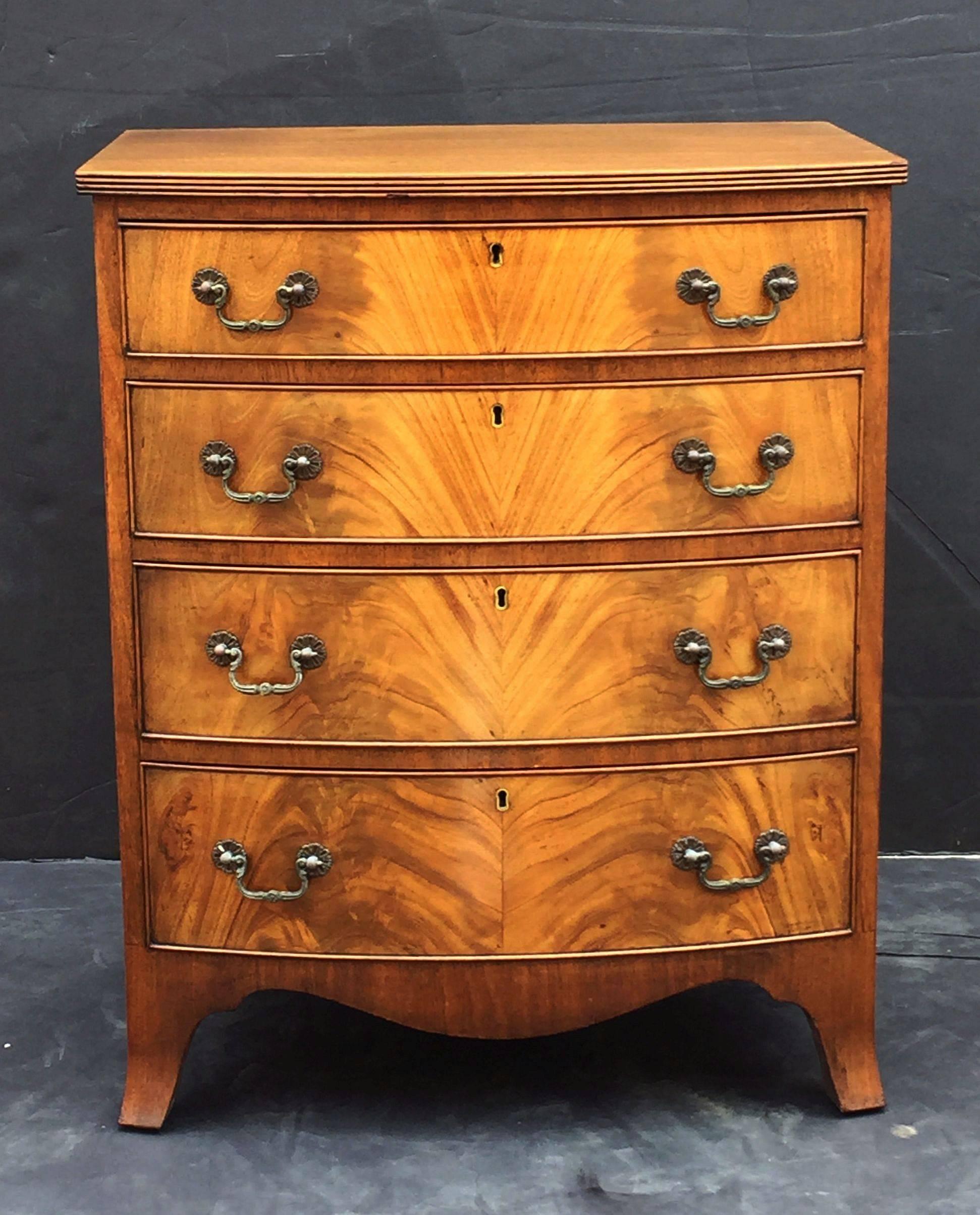 A fine English small bow front chest of mahogany, featuring a moulded top over a frieze of four bowed and beaded drawers, each drawer with handsome flame-cut mahogany veneers and brass pulls and escutcheons. 
Set upon a serpentine apron base with