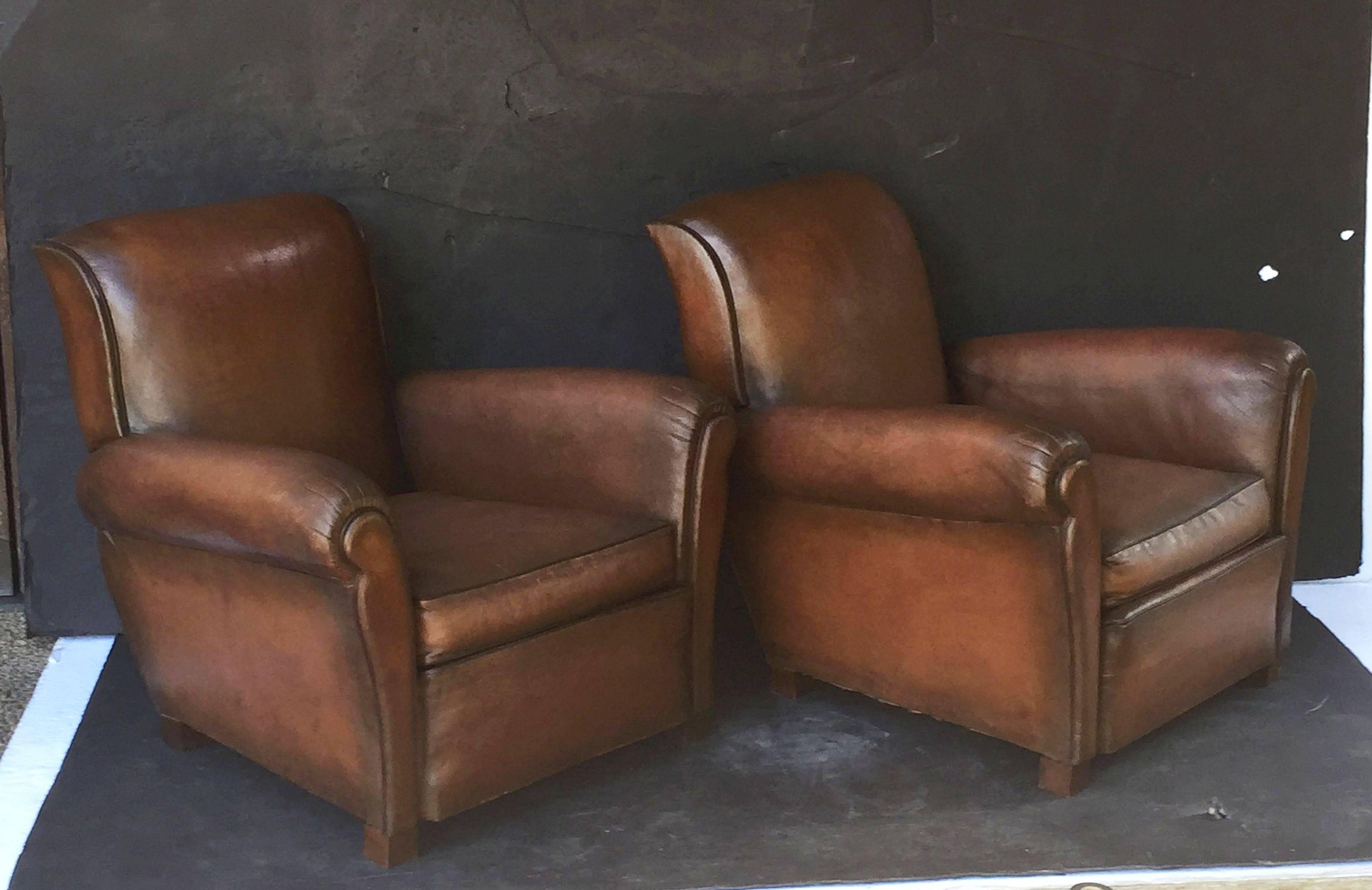 A handsome pair of vintage French leather upholstered club or lounge chairs, each chair featuring a comfortable back and seat with removable cushion, with stylish arms and original leather, and brass nailhead trim to back. Resting on block