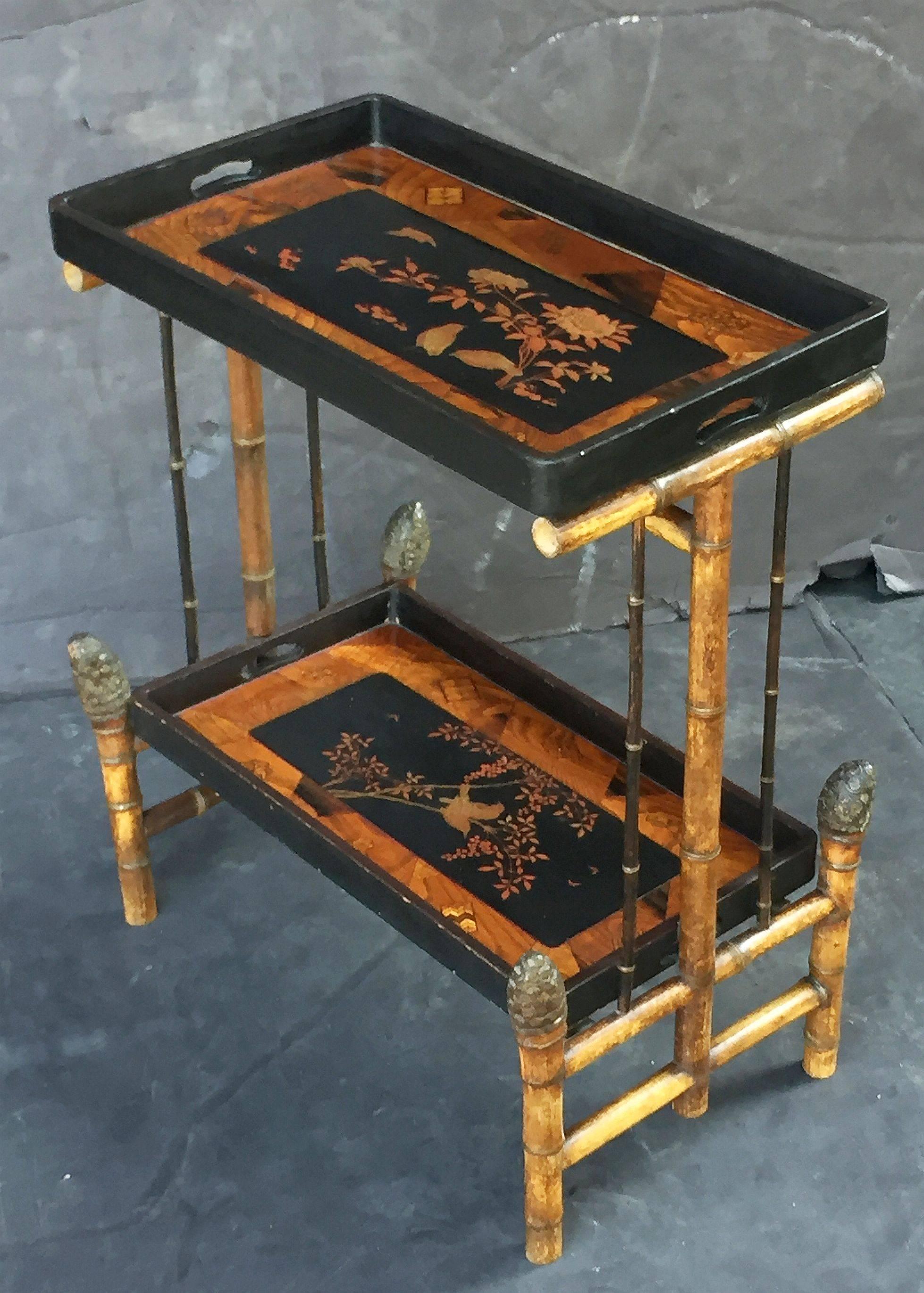 A fine English bamboo rectangular tray table featuring two tiers of ebonized and lacquered panel trays, each tray with marquetry accents around a chinoiserie design in the center, mounted to a stylish frame of bamboo, with bamboo root foot finials