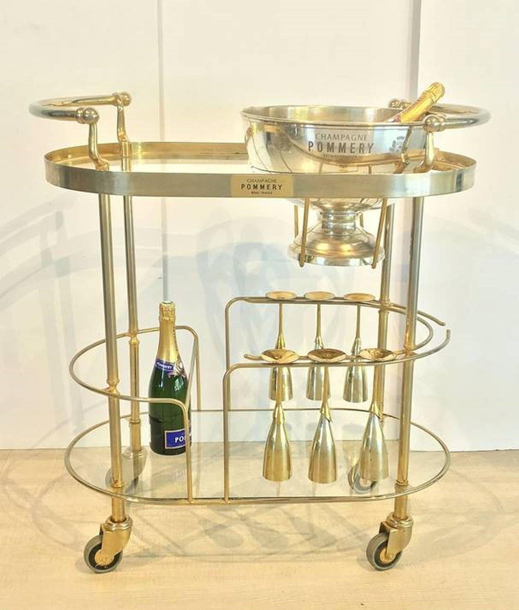 An Art Deco champagne trolley or rolling bar cart
featuring a two-tiered curved frame of chromed brass and glass,
the top tier with fitted silver bowl or wine cooler for ice,
the second tier with six champagne flutes of silver and brass,
on vintage