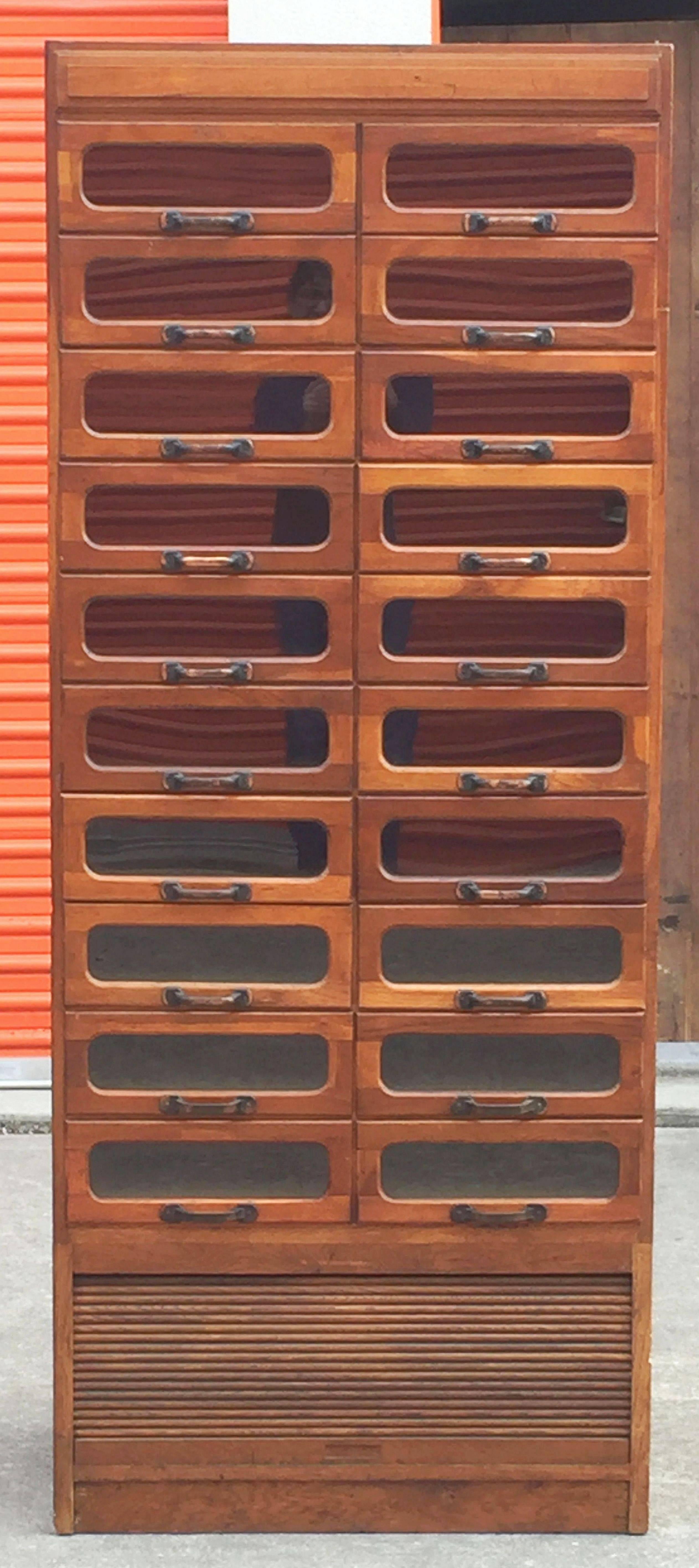 An exceptional large haberdashery or haberdasher's cabinet from England featuring:

(20) glass-fronted drawers over (one) blind rolltop tambour shutter cupboard - (21) drawers total.

The fitted drawers with metal hardware and set upon a plinth